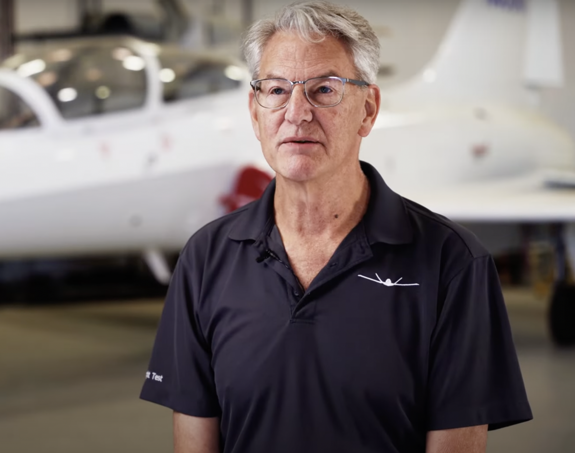 An older man with gray hair and glasses, wearing a black polo shirt, standing in front of a white aircraft inside a hangar.