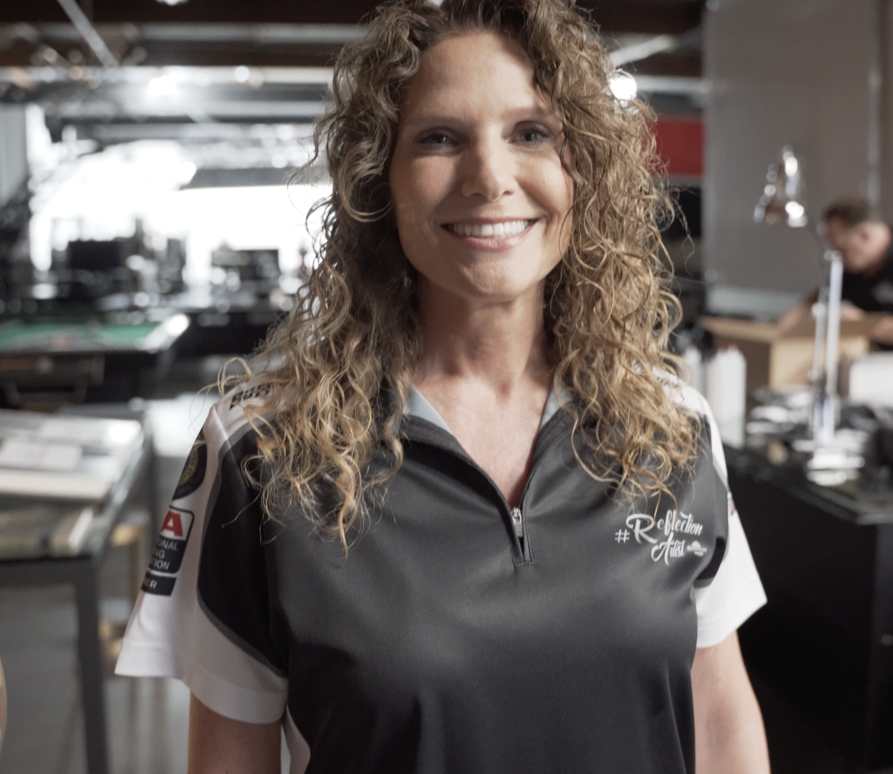 A woman with curly hair smiling in a casual setting, wearing a sports polo shirt with patches and writing on the front, possibly in a workshop or studio environment.