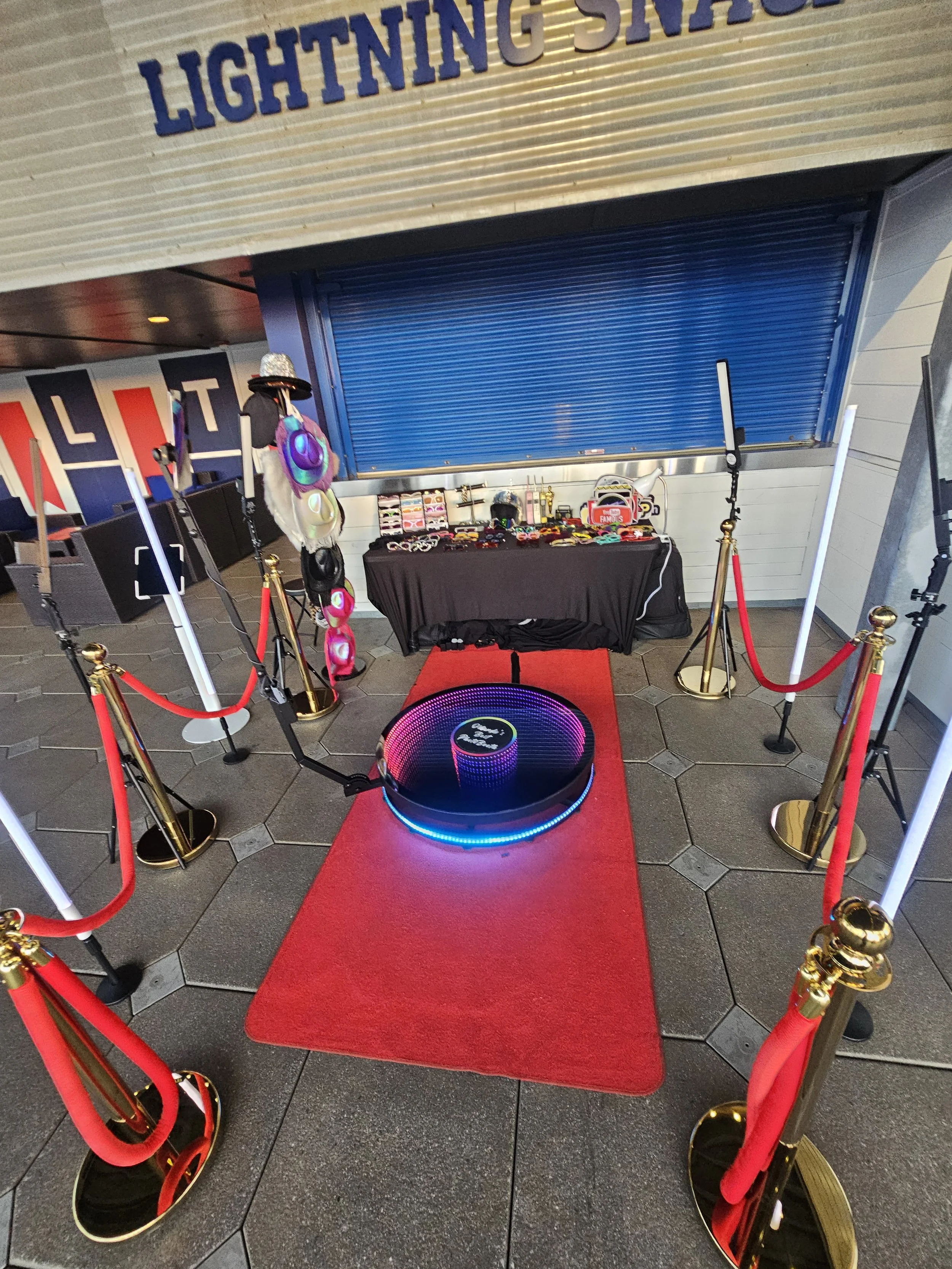 A virtual reality photo booth setup with a red carpet, gold stanchions, red ropes, a purple-lit VR headset, a table with accessories and merchandise, and a display of hats and headphones in the background, under a sign that says "Lighting SNACK".