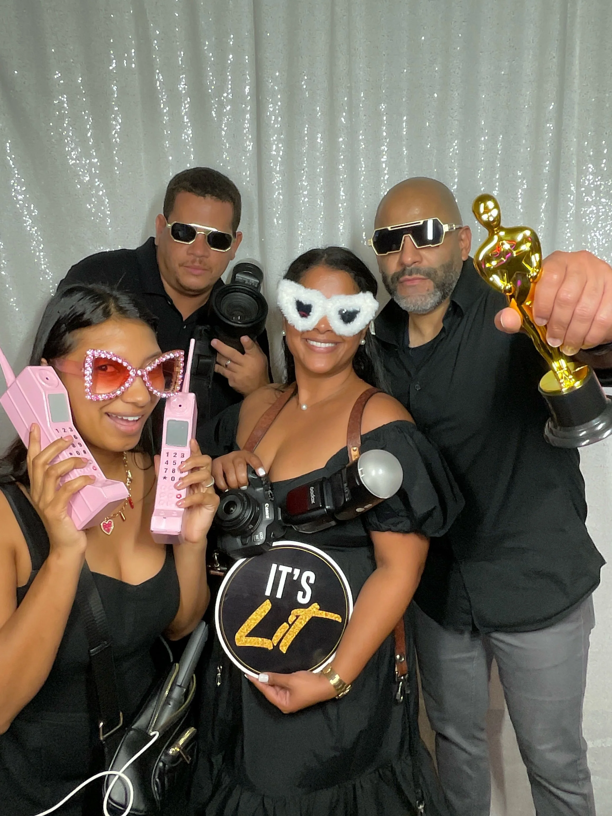 Group of five people at a celebration, all wearing fun accessories like sunglasses, masks, and holding awards, posing in front of a sparkly backdrop.