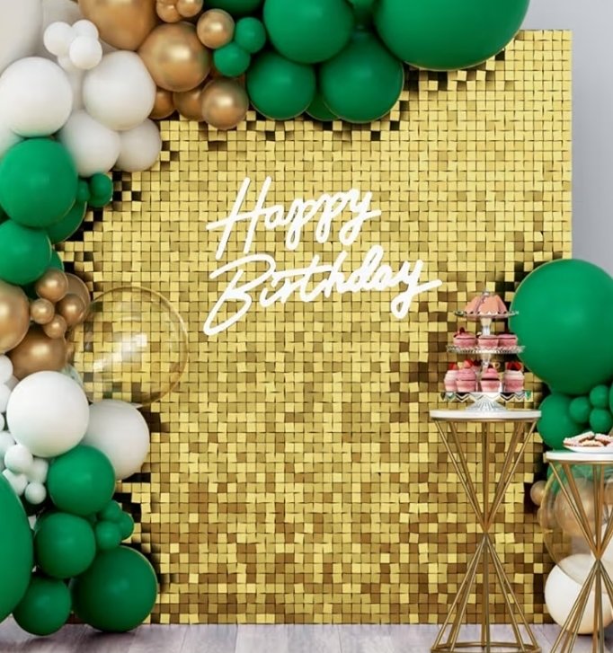Gold mosaic backdrop with white neon sign that says "Happy Birthday" surrounded by green, white, and gold balloons. Pink cupcake stand with cupcakes on the right and a small round gold table with snacks.