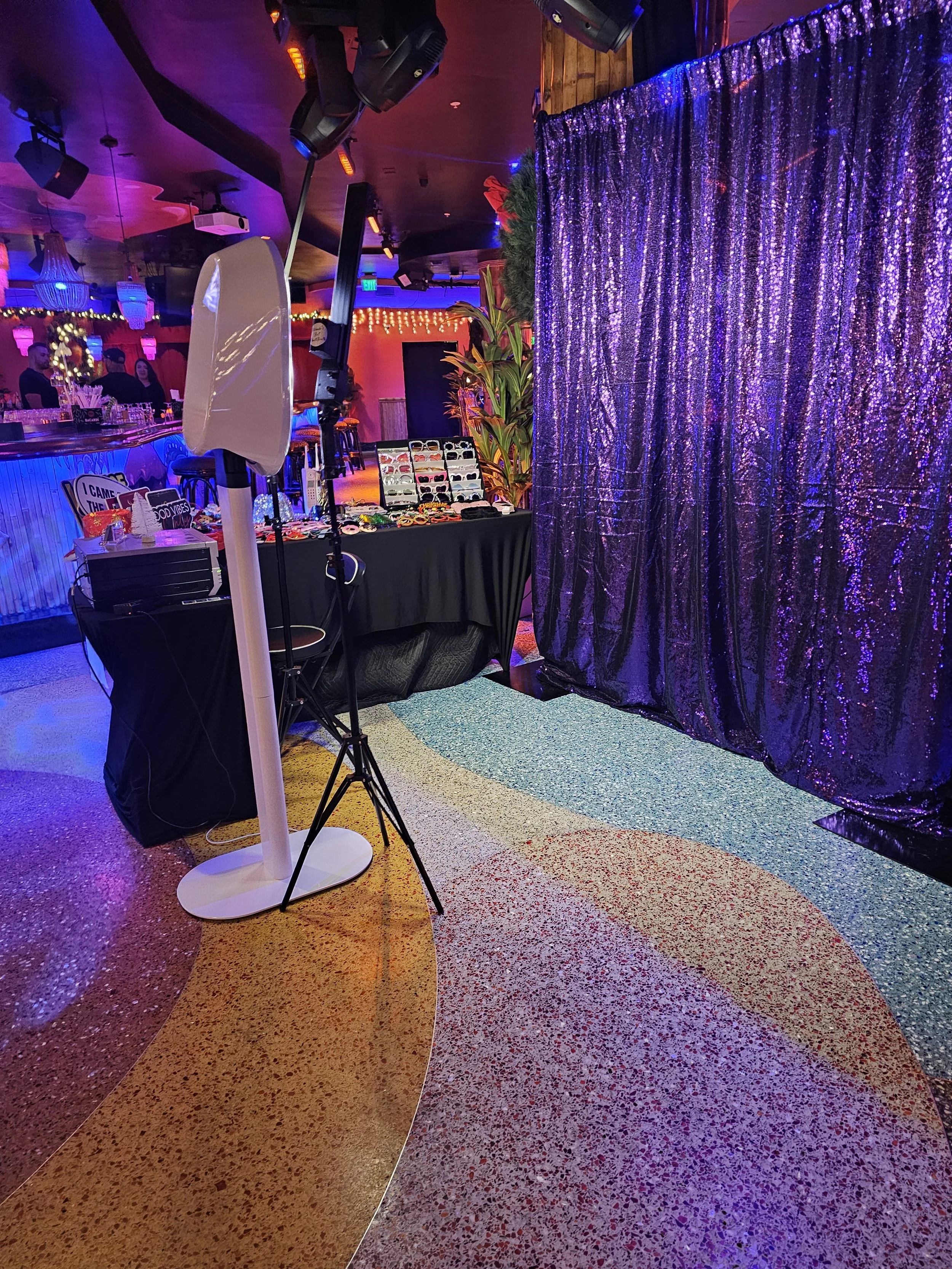 Photo of an indoor event space with a colorful, patterned floor, a purple glittery curtain, and a table with eyewear products and accessories. There is a lighting stand and a black tablecloth, with a lively, festive atmosphere.