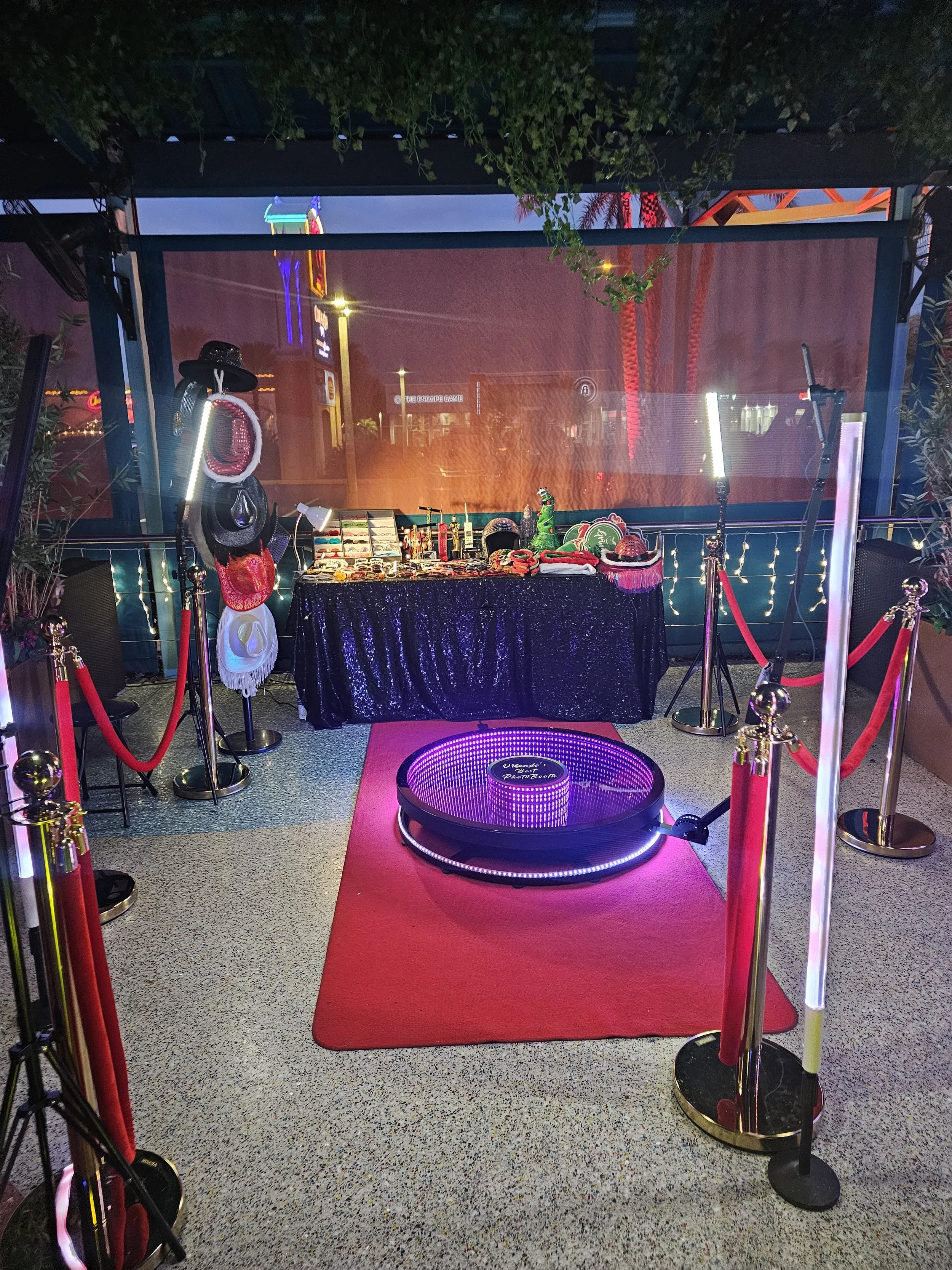 Photo of a photo booth setup at night with a starry lighted circle and a table with photo props, accessories, and decorations. Red and gold stanchions with ropes gather around the area. There is a dark cloth on the table, and bags and hats hang on a stand to the left. String lights and colorful neon signs are visible in the background.