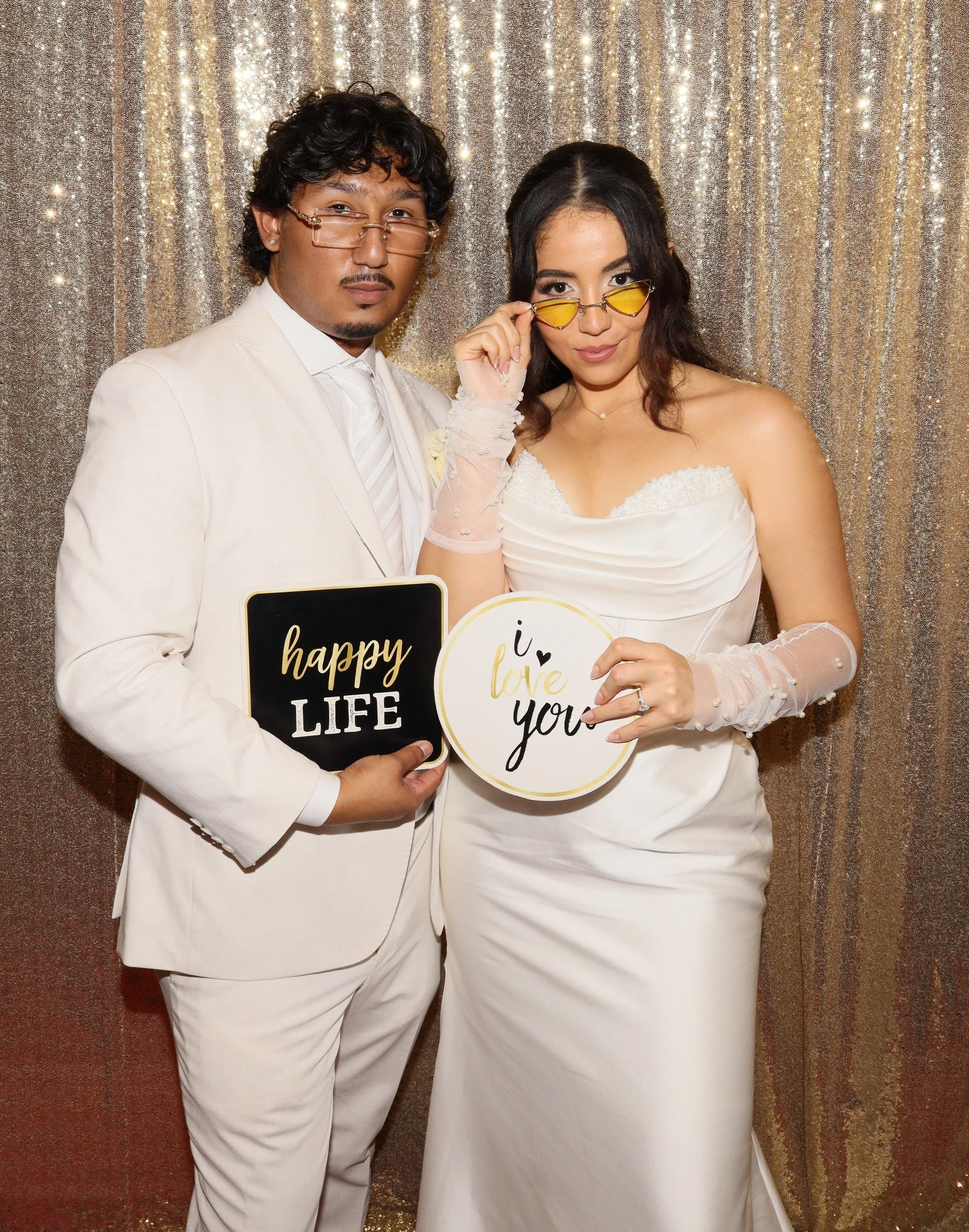 A man and woman dressed in white tuxedo and wedding dress pose in front of a sparkly gold backdrop, holding signs that say 'happy life' and 'i love you'.