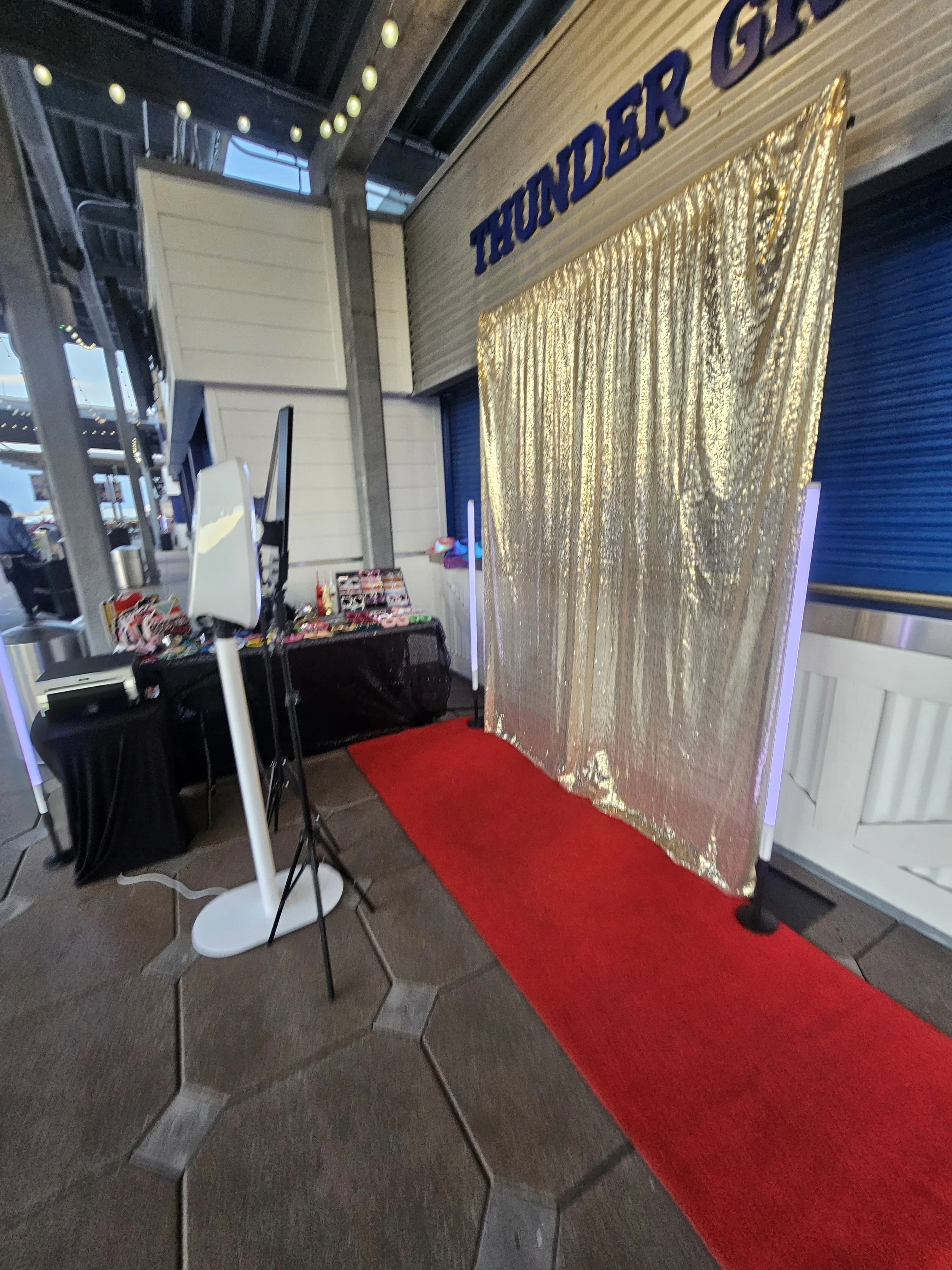 Photo of an event entrance with a red carpet leading to a gold shimmering curtain, above which the word 'THUNDER' is visible. To the left, there are tables with promotional items and photo booths.