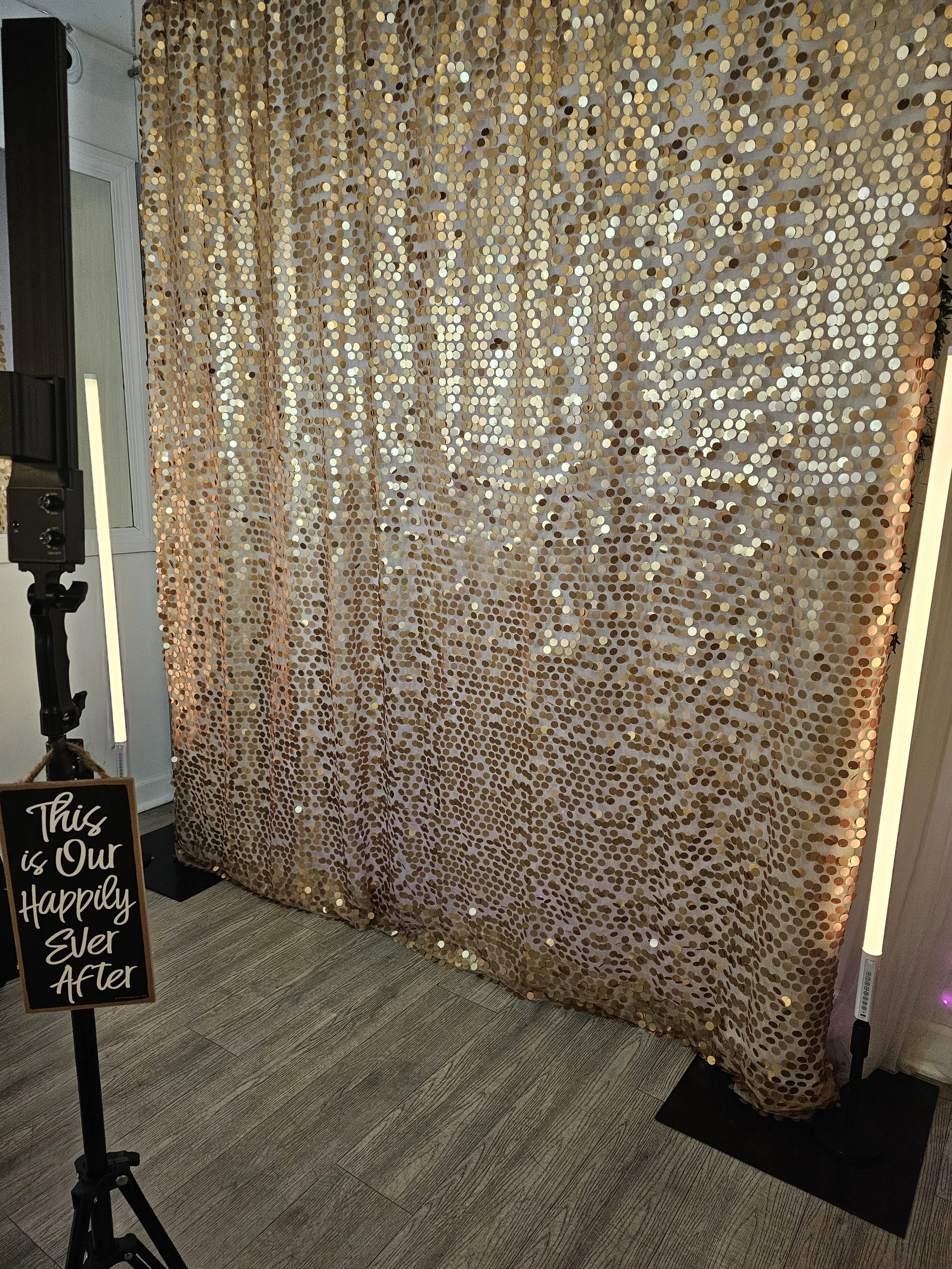 A sparkling gold sequin curtain backdrop with a sign that reads "This is Our Happily Ever After" on a small black easel in front of the curtain, set up for a celebration or wedding event.