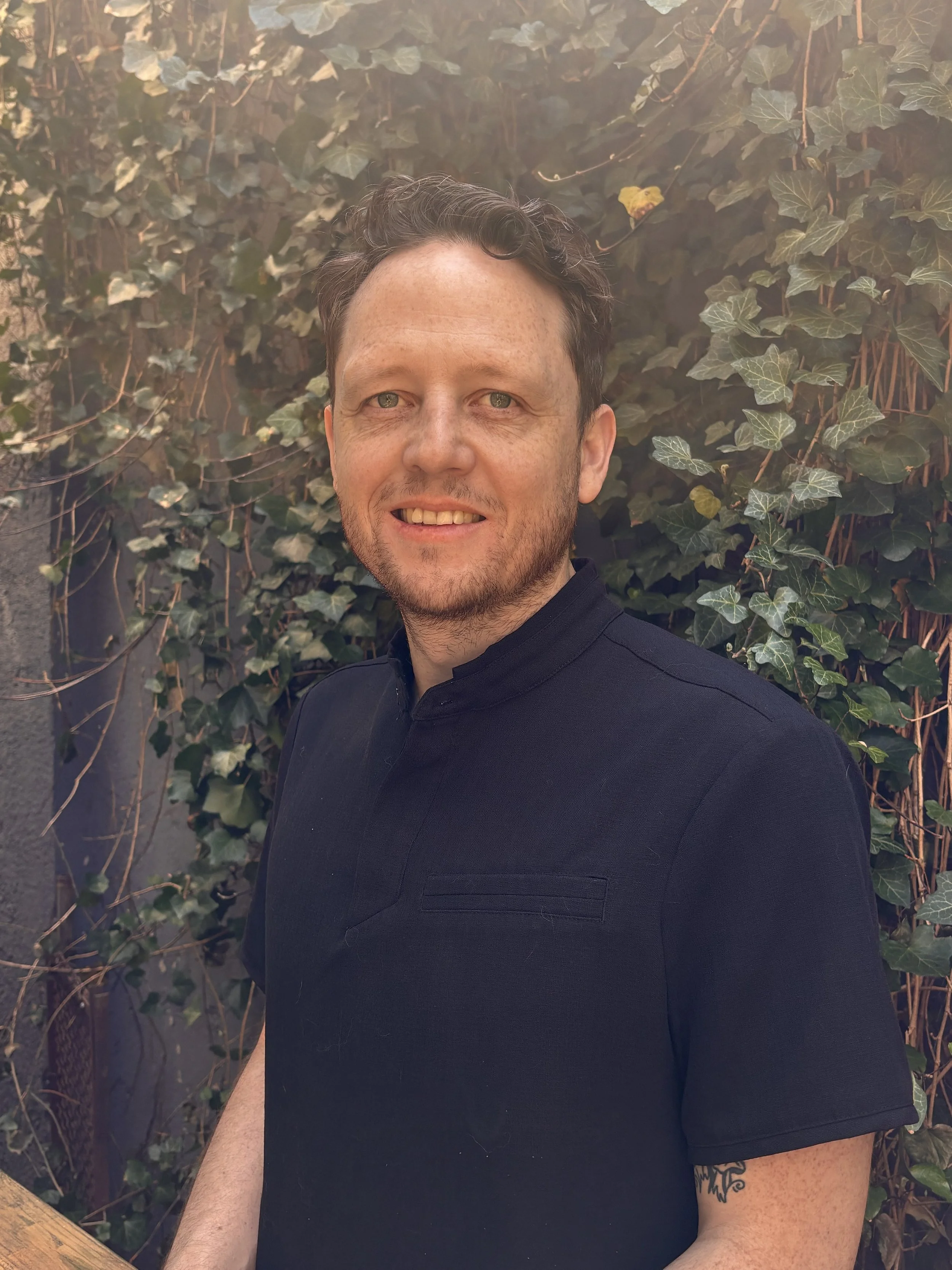 Gareth, massage therapist, smiles while standing outdoors wearing a dark shirt with greenery in the background.