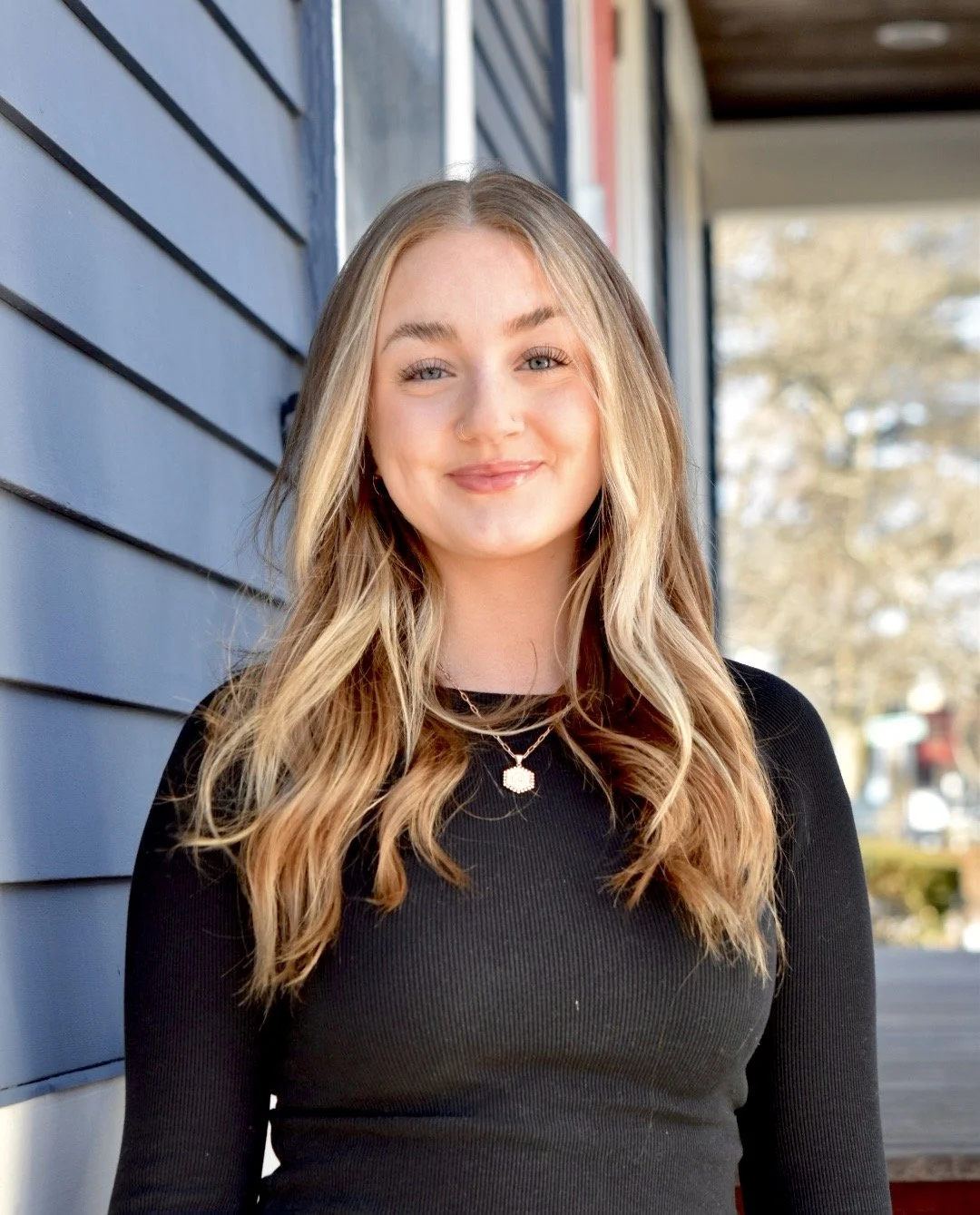 Grace C., esthetician specializing in skincare, facials, and waxing, smiles while standing outdoors in front of a blue building, wearing a black top.