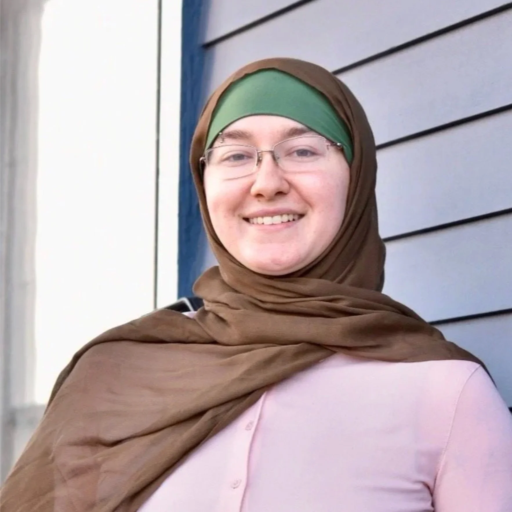Autumn L., massage therapist, smiles while standing outdoors wearing glasses, a brown shawl, and a patterned scarf.
