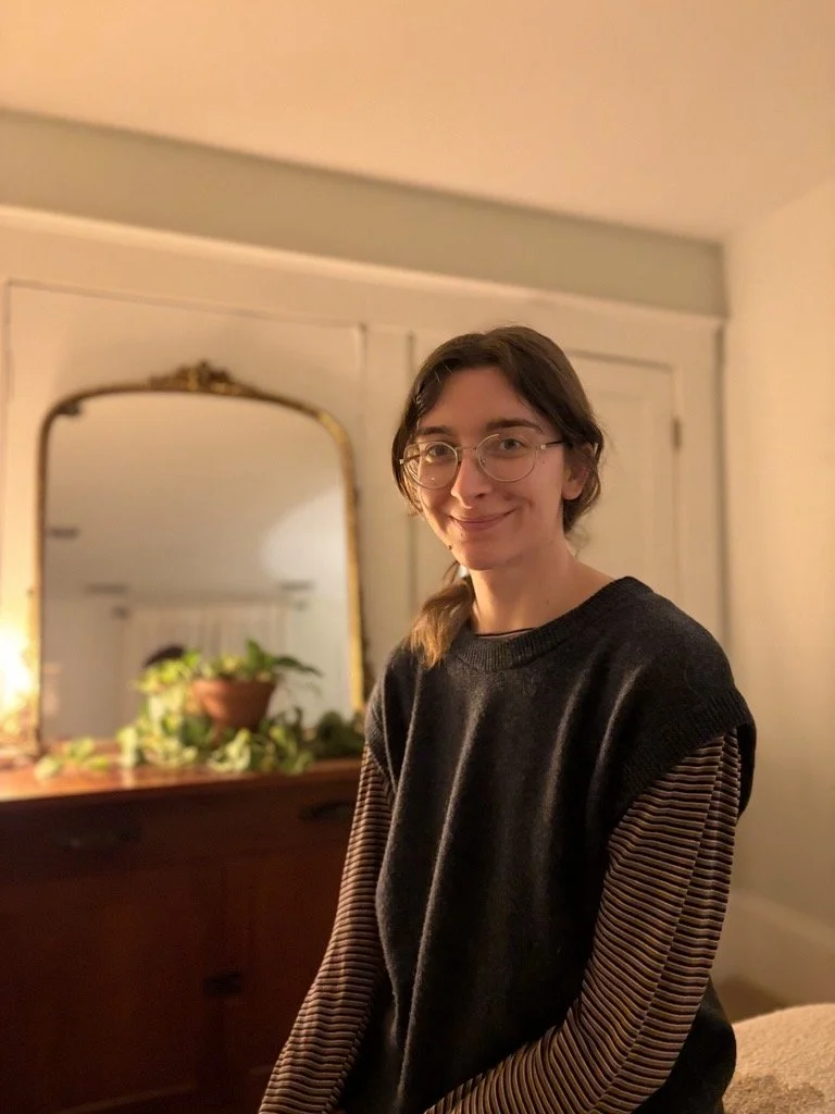 Abbey N., massage therapist, smiles while standing indoors wearing a black top in a warmly lit treatment space.