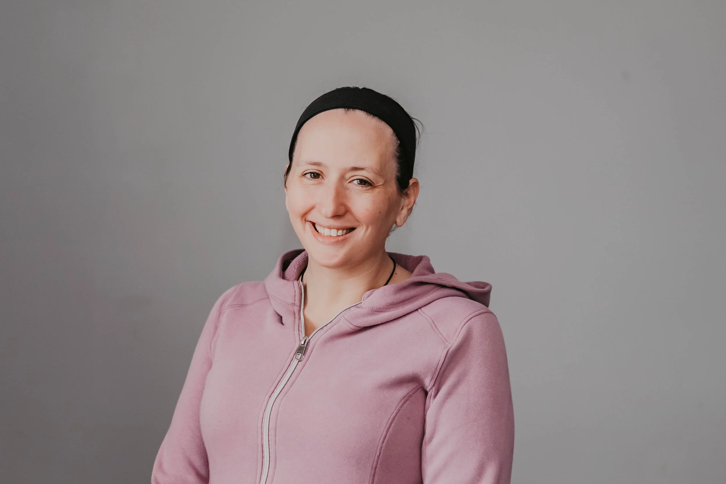 Nicole N., master massage therapist specializing in hot stone massage and prenatal massage, smiles while standing against a light gray background wearing a pink hoodie.