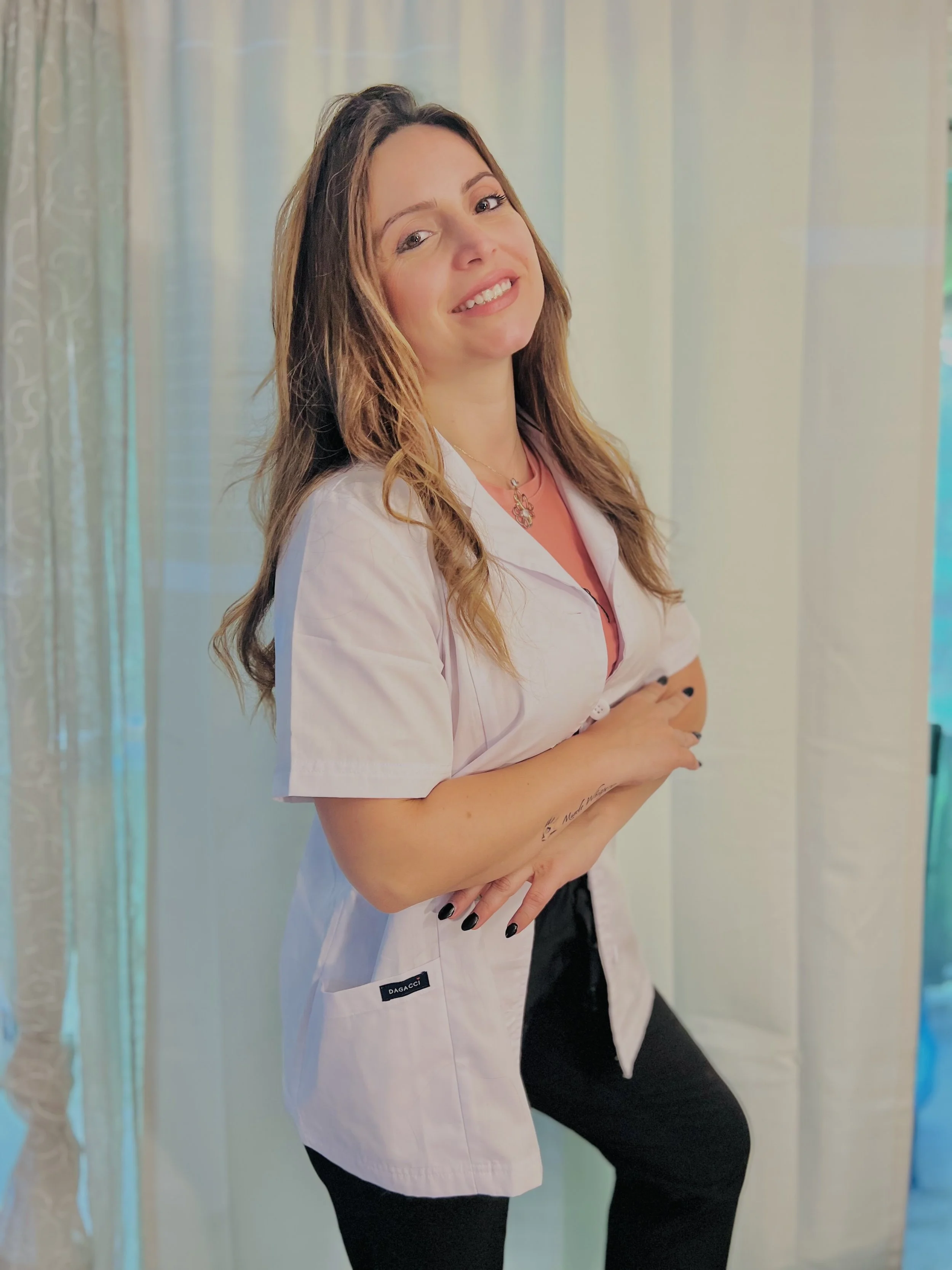 Graziella S., massage therapist, smiles while standing indoors wearing a white therapist uniform with her hands folded in front of her.