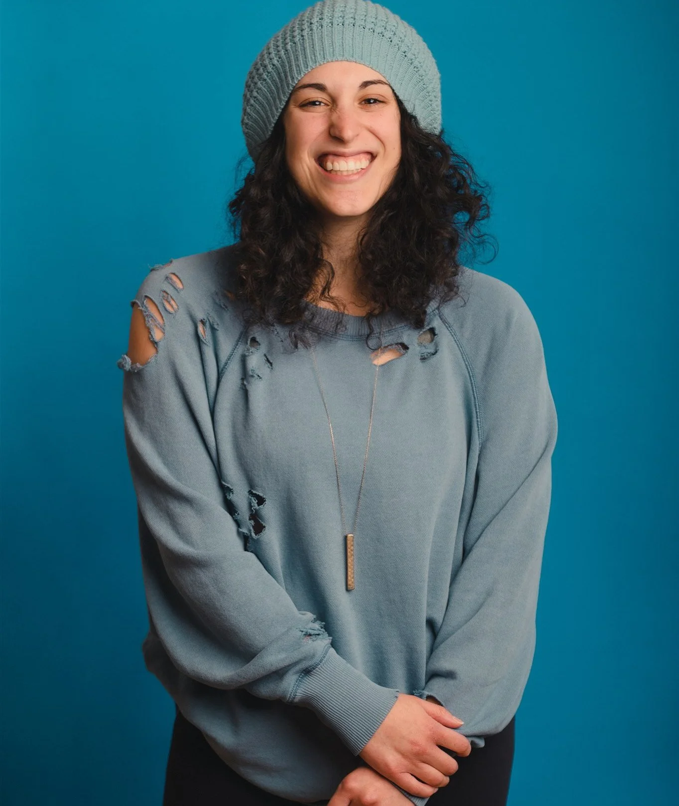 Mallory, desk concierge, smiles while seated against a blue background wearing a gray sweater and knit beanie.