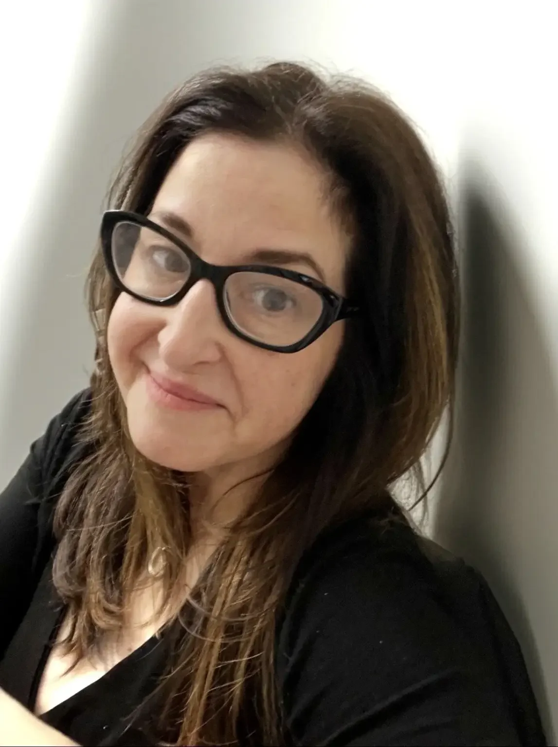 Mandalynn R., Esthetician, smiles toward the camera. She has long dark brown hair, is wearing black-framed glasses and a black top, and is leaning slightly toward the camera.