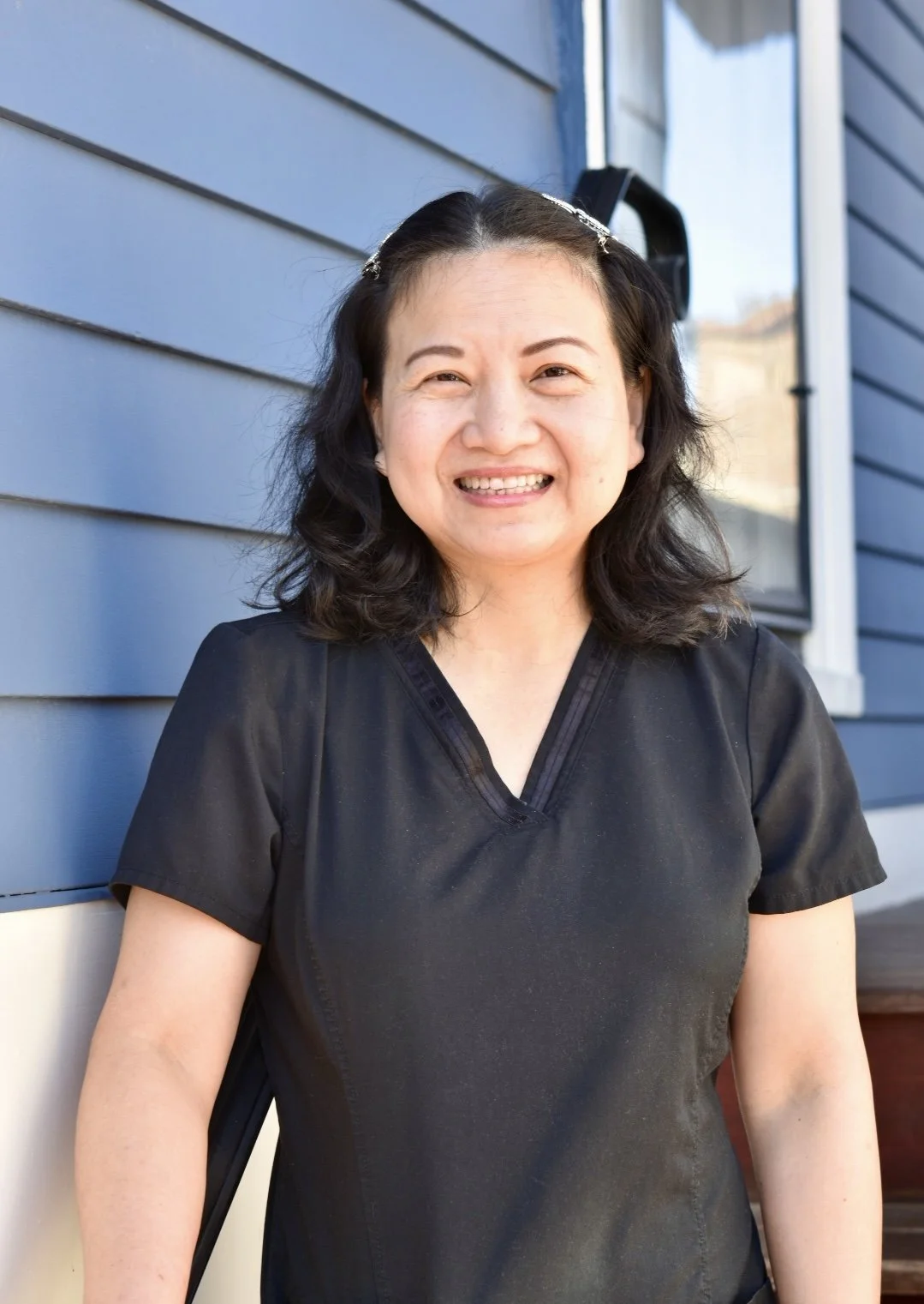 Bella K., master massage therapist, smiles while standing outdoors in front of a blue building wearing a black sweater.