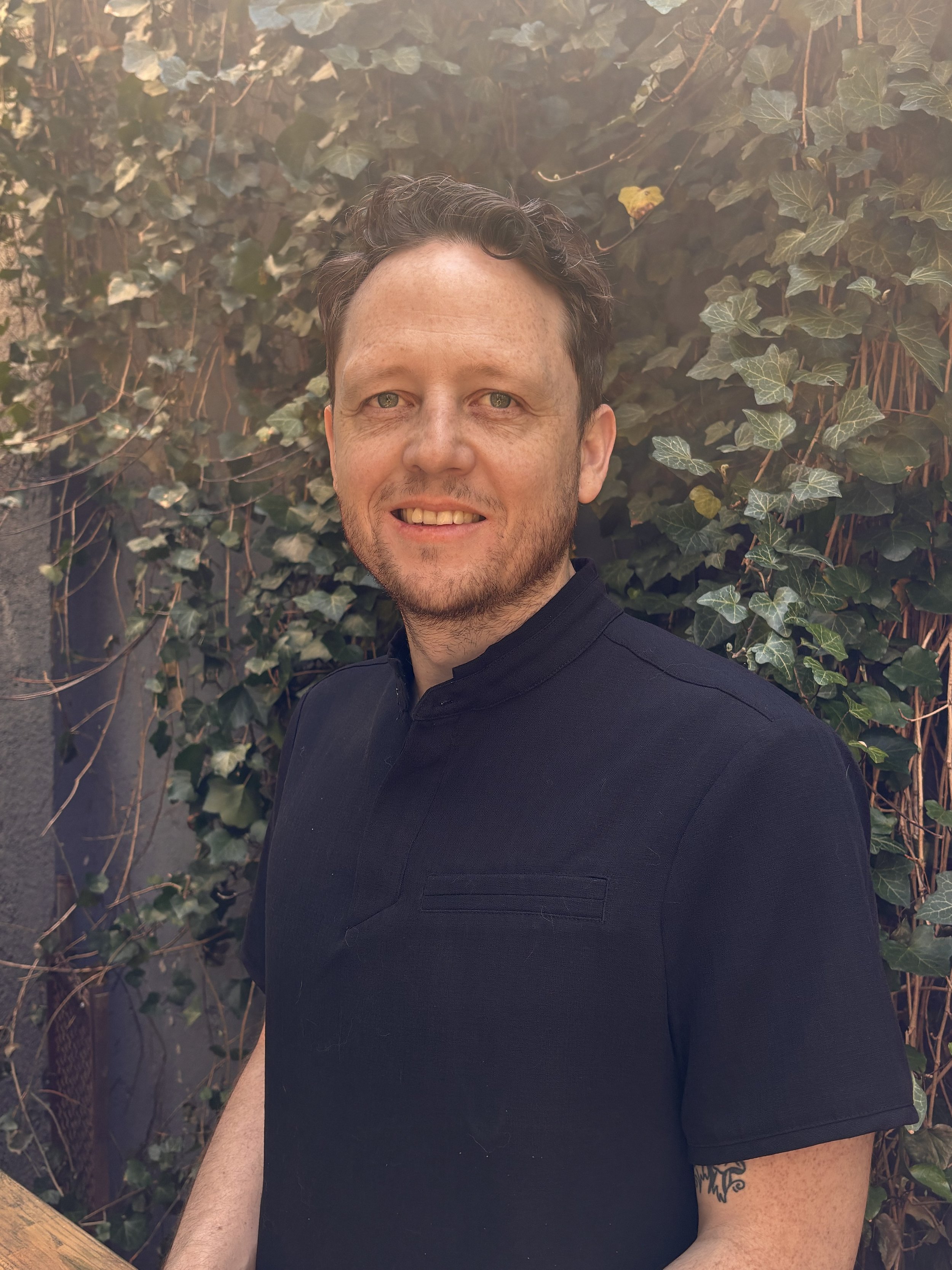 Gareth, massage therapist, smiles while standing outdoors wearing a dark shirt with greenery in the background.