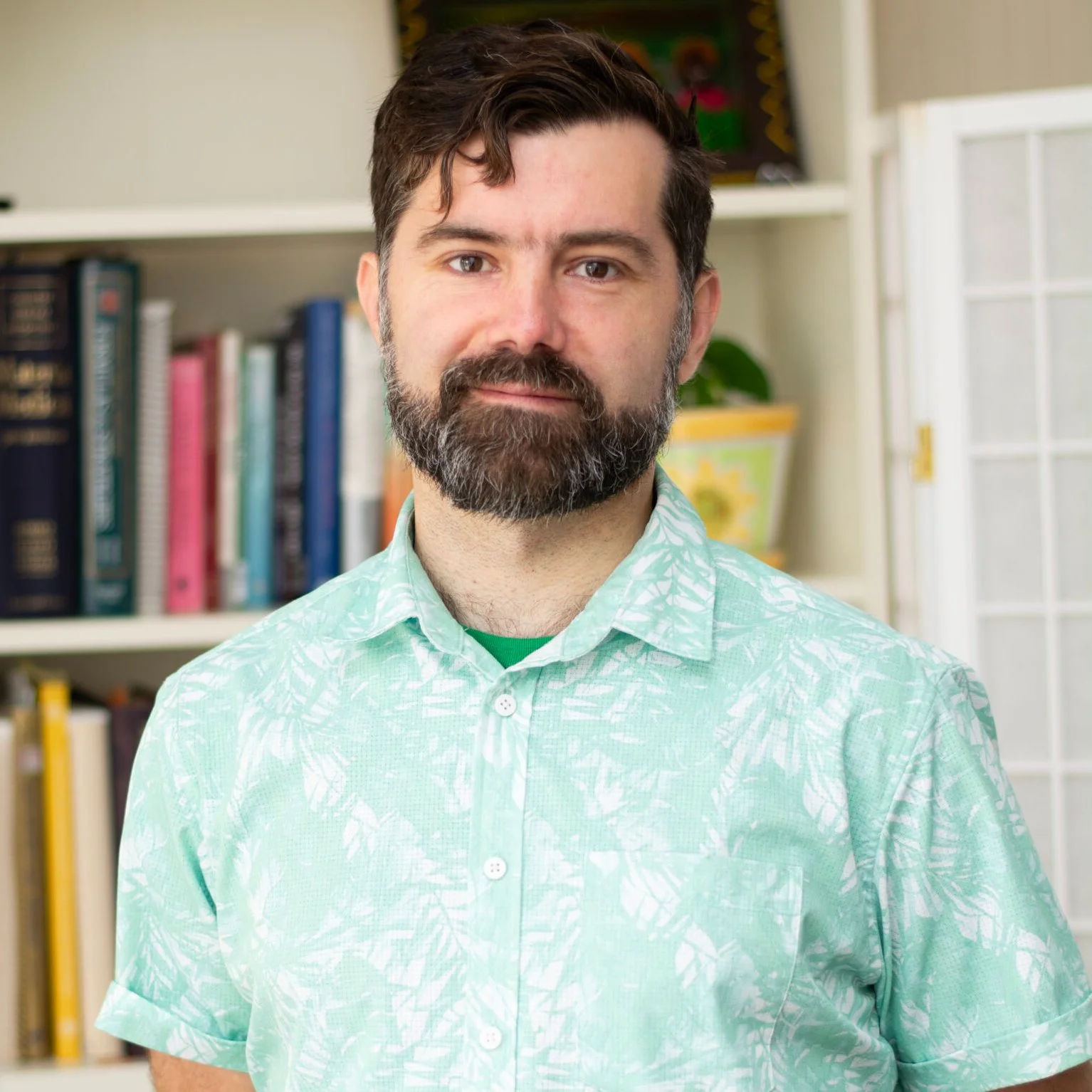 Andrew C., licensed acupuncturist, smiles while standing indoors wearing a light green patterned shirt with bookshelves in the background.