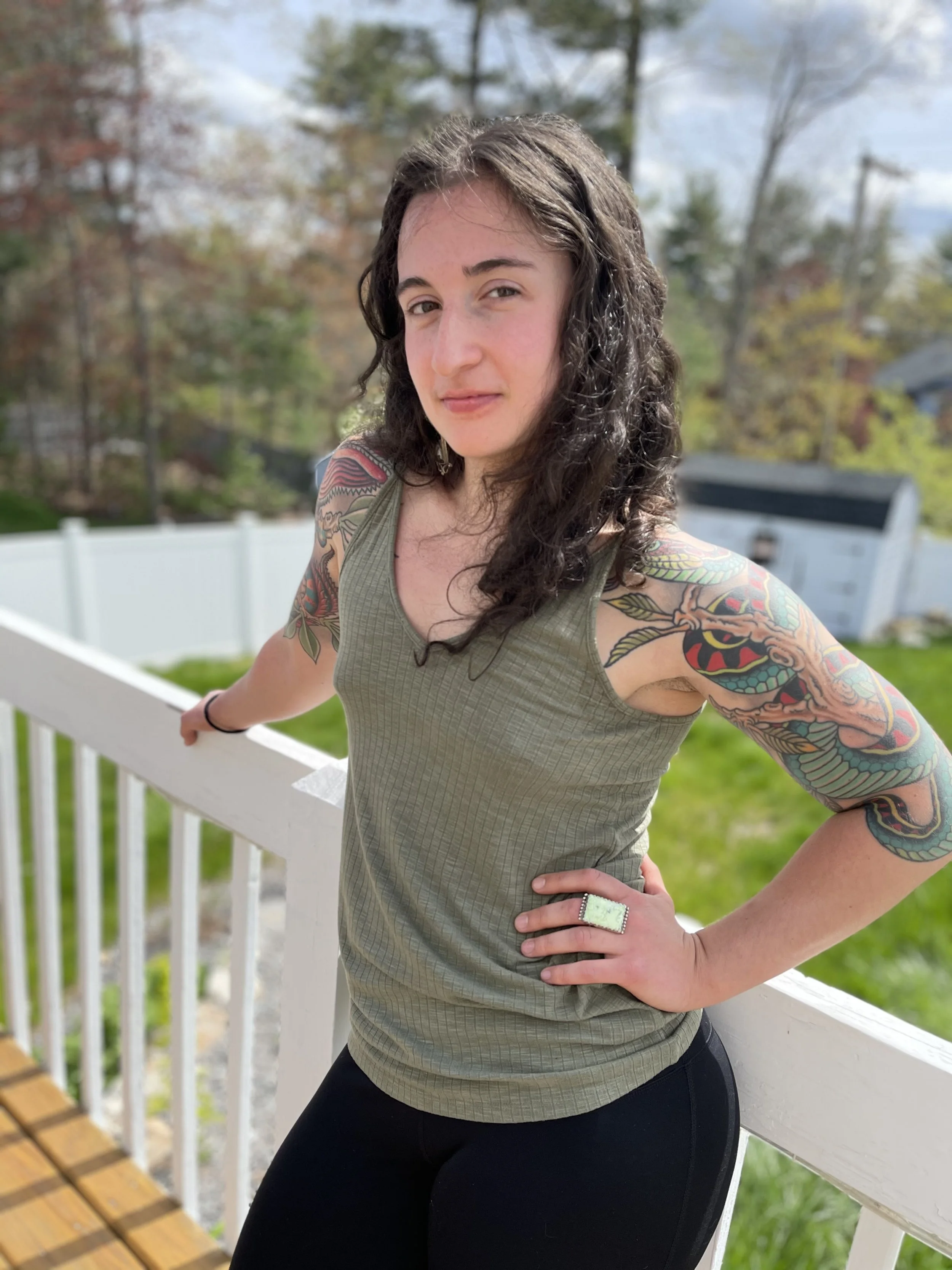This image of Claire is taken outside on a porch. Claire is wearing a green tank top and has tattoo art. Her hair is down. She is looking directly at the camera and standing with one hand on one hip and the other hand on the porch railing.