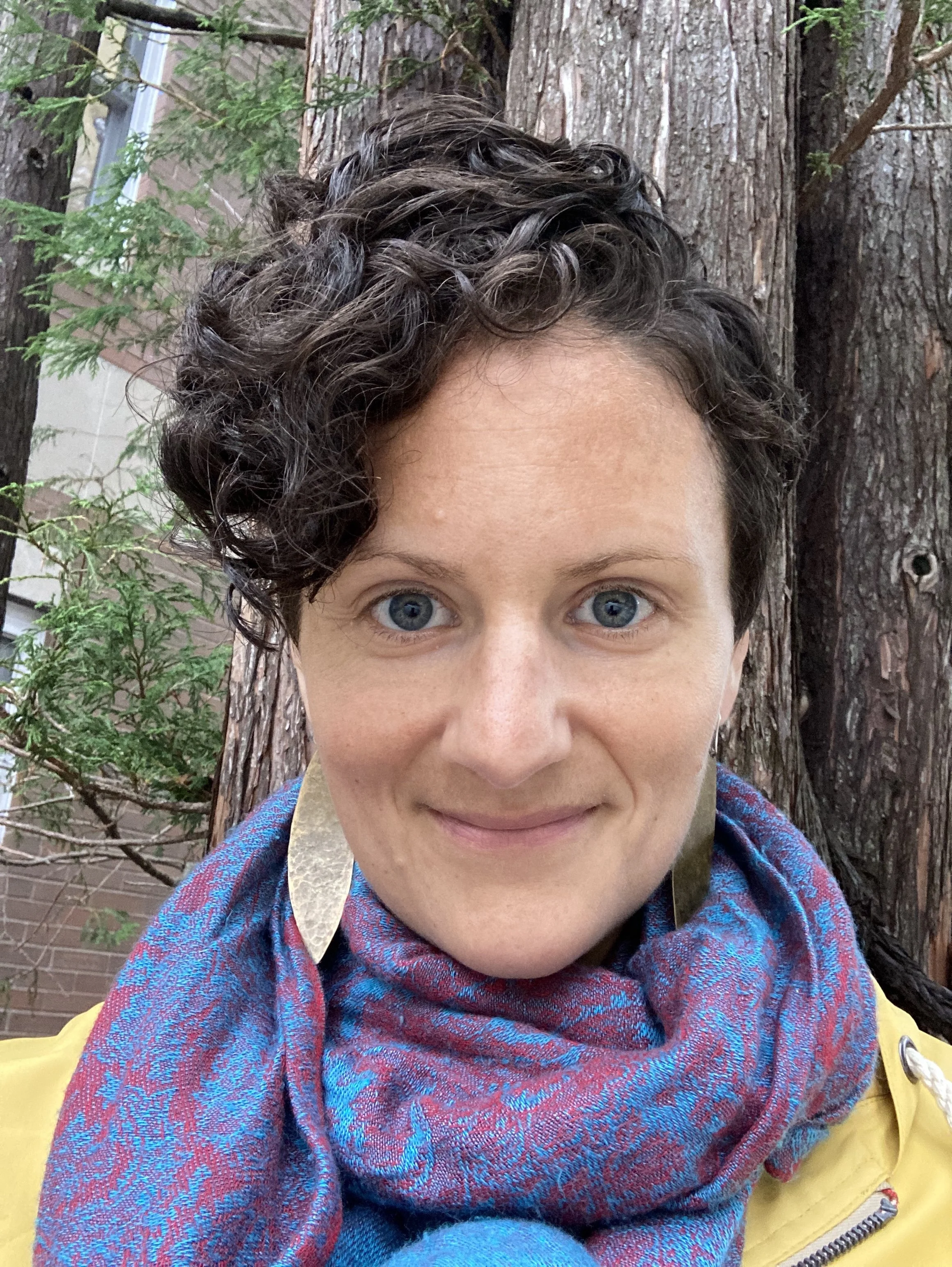 Lisa D., sound bath practitioner, smiles while standing outdoors wearing a colorful blue and purple scarf.