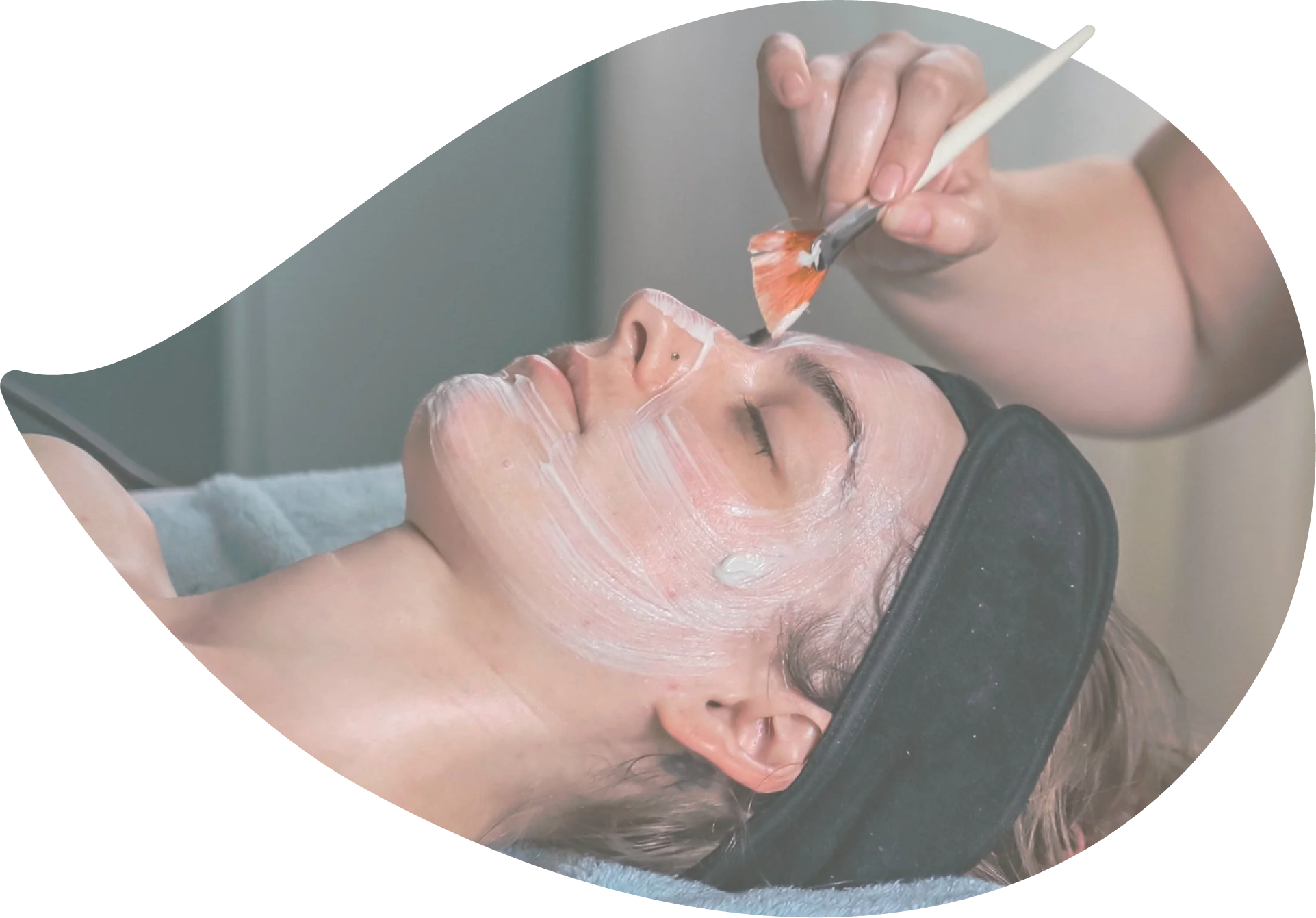 A client relaxes with their eyes closed while a practitioner applies a facial treatment mask using a soft brush.