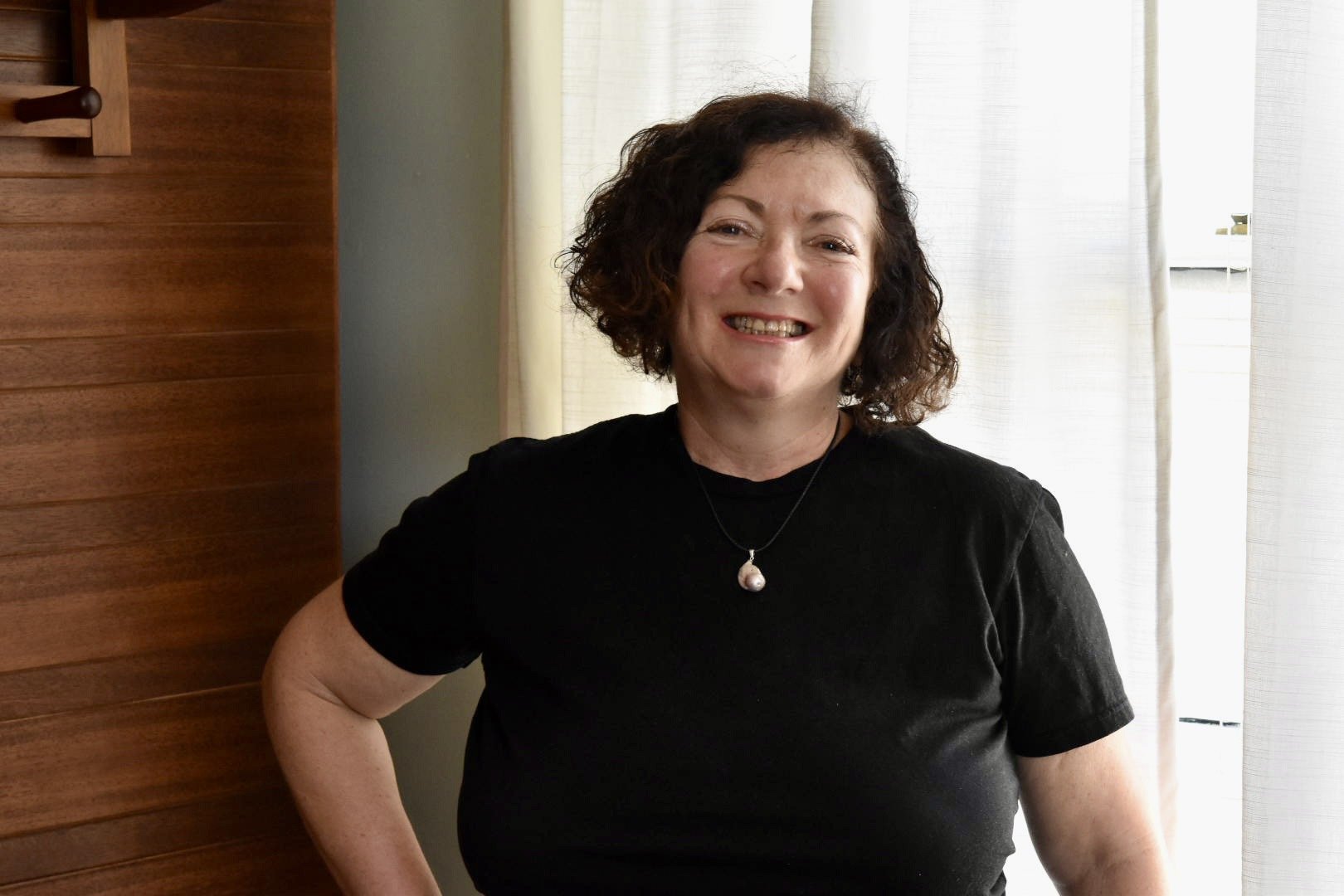 Anna C., master massage therapist, smiles while standing indoors wearing a black top against a light-colored wall.