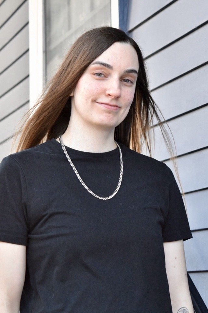 Quinn S., massage therapist, smiles while standing in front of a blue building wearing a black shirt.