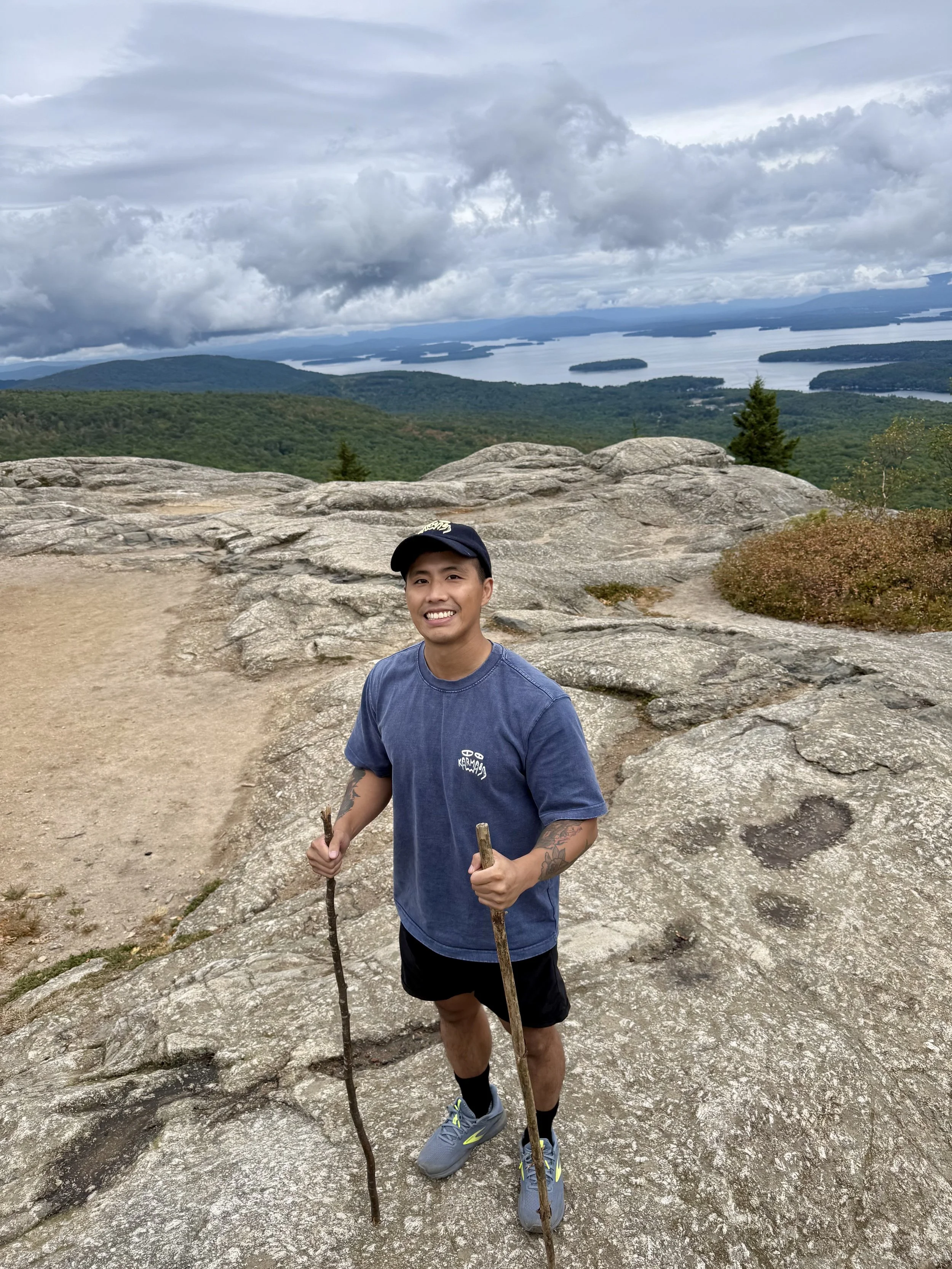 Mitchell M., massage therapist, smiles while standing outdoors on a rocky hillside wearing a blue shirt.