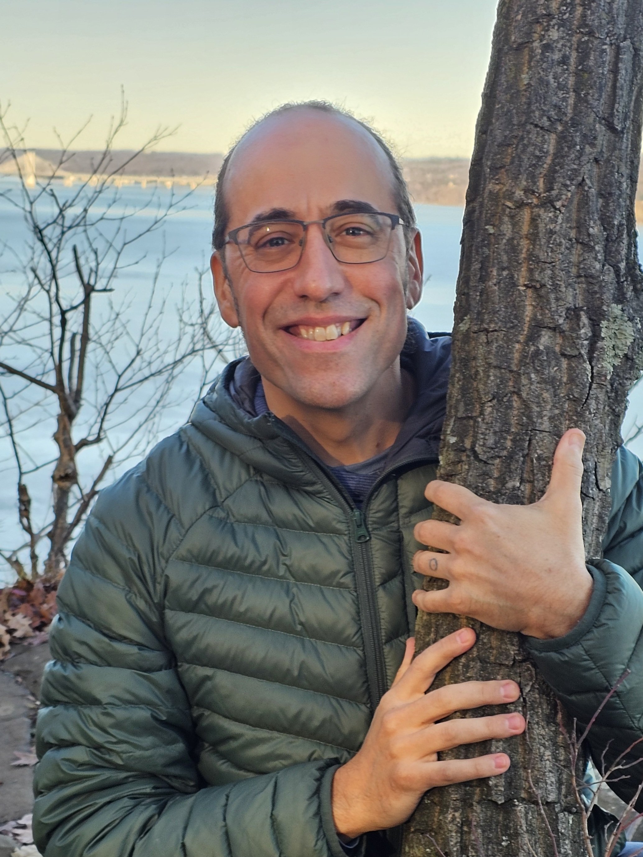 Nelson T., master massage therapist specializing in myofascial release, smiles while standing outdoors beside a tree, wearing a green jacket over a blue shirt.