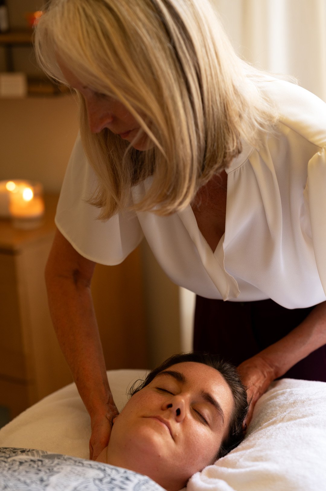 A woman receives a massage from Julie in a cozy, softly lit room with a lit candle in the background.