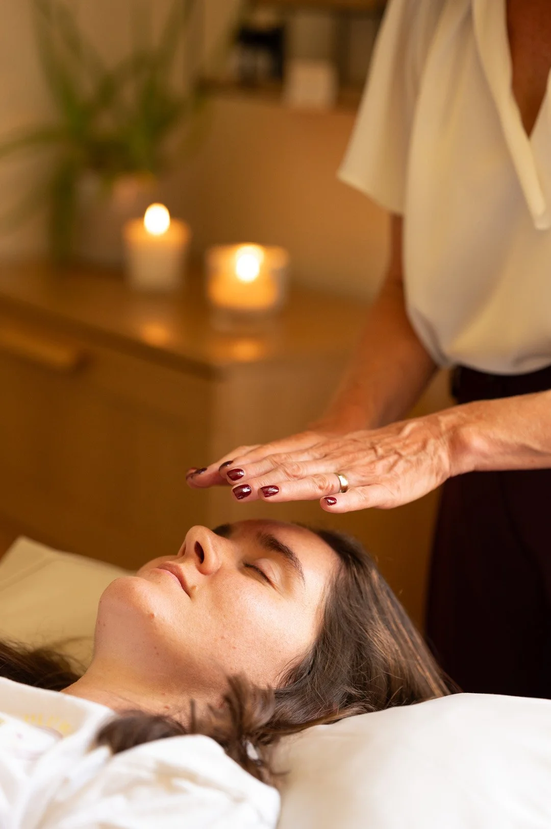 A woman receiving a facial massage in a spa setting with lit candles and a blurred plant in the background.