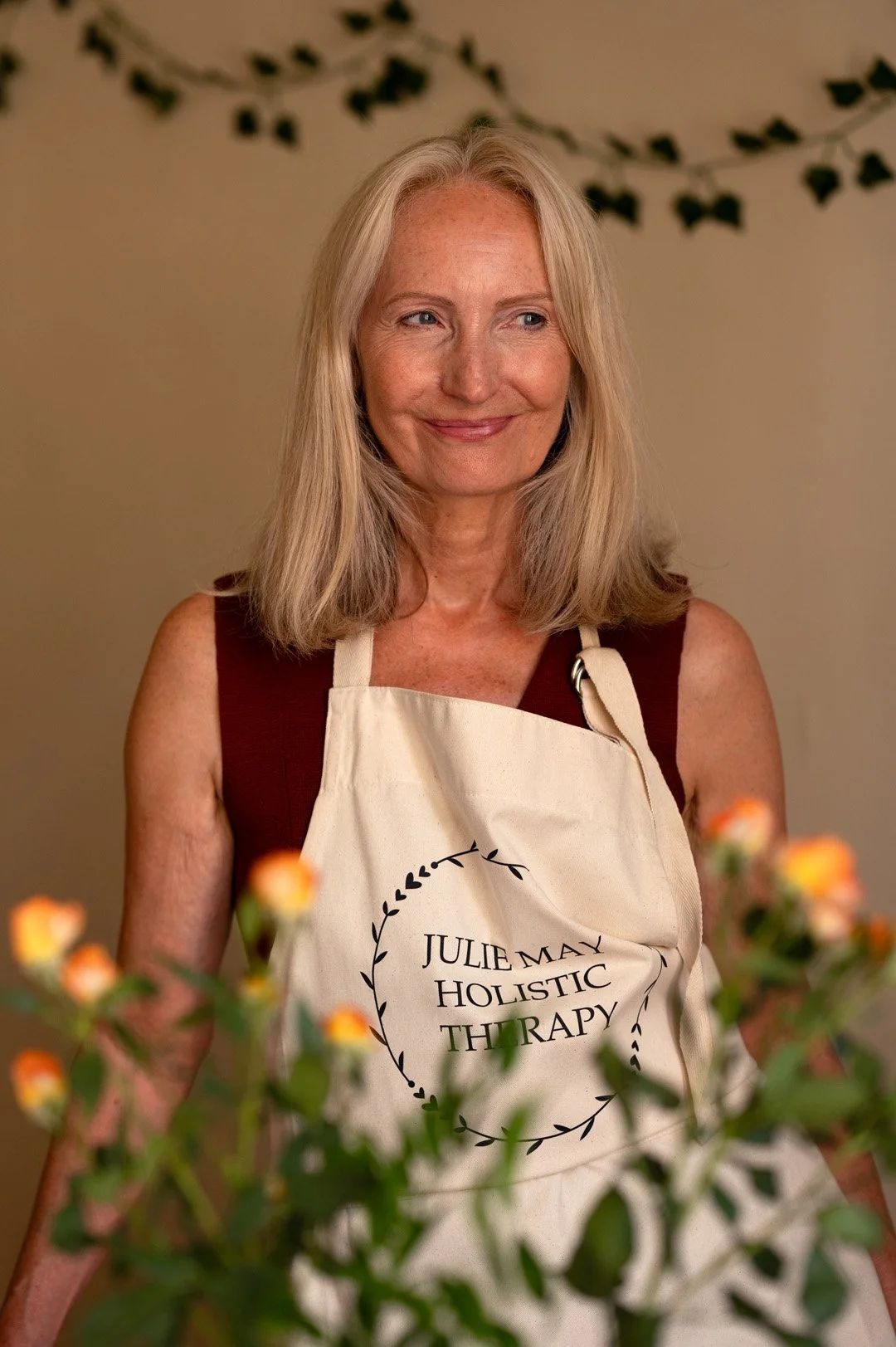 A smiling woman with blonde hair wearing a beige apron that reads 'Julie May Holistic Therapy,' standing behind flowers with a blurred background.