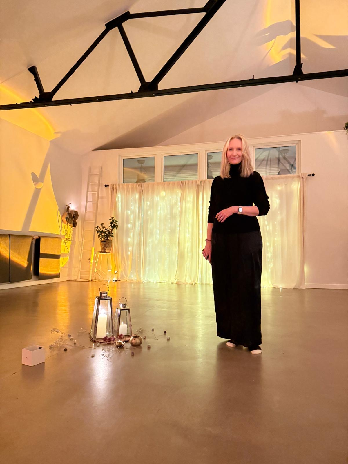 Julie standing in a decorated indoor event space in Bristol with warm lighting, string lights, a curtain backdrop, and candles on the floor for a holistic event.