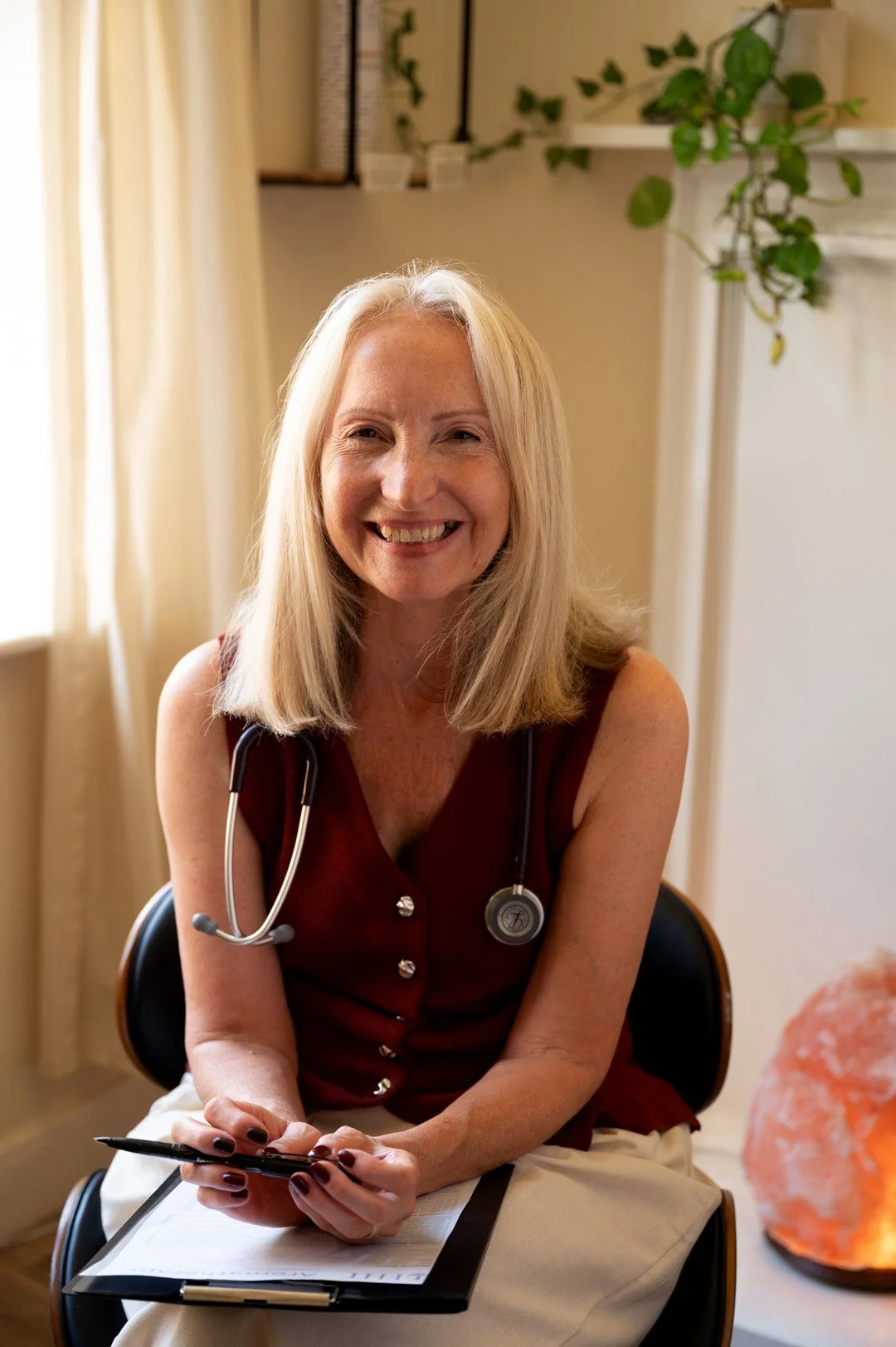 Julie wearing a sleeveless maroon top, sitting at a desk with a clipboard and pen, and a stethoscope around her neck in a well-lit room with beige curtains and green plants.