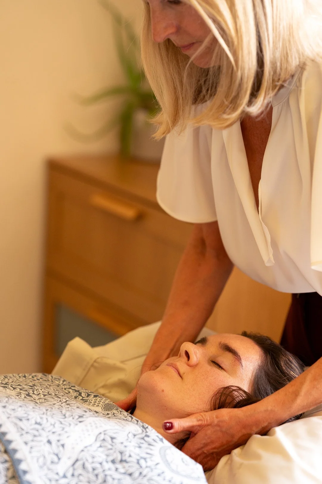 A woman is receiving a massage while lying on a bed or massage table, with her eyes closed and a relaxed expression. The massage is being performed by a person with blonde hair, wearing a white shirt. There is a wooden furniture piece and a green plant in the background.