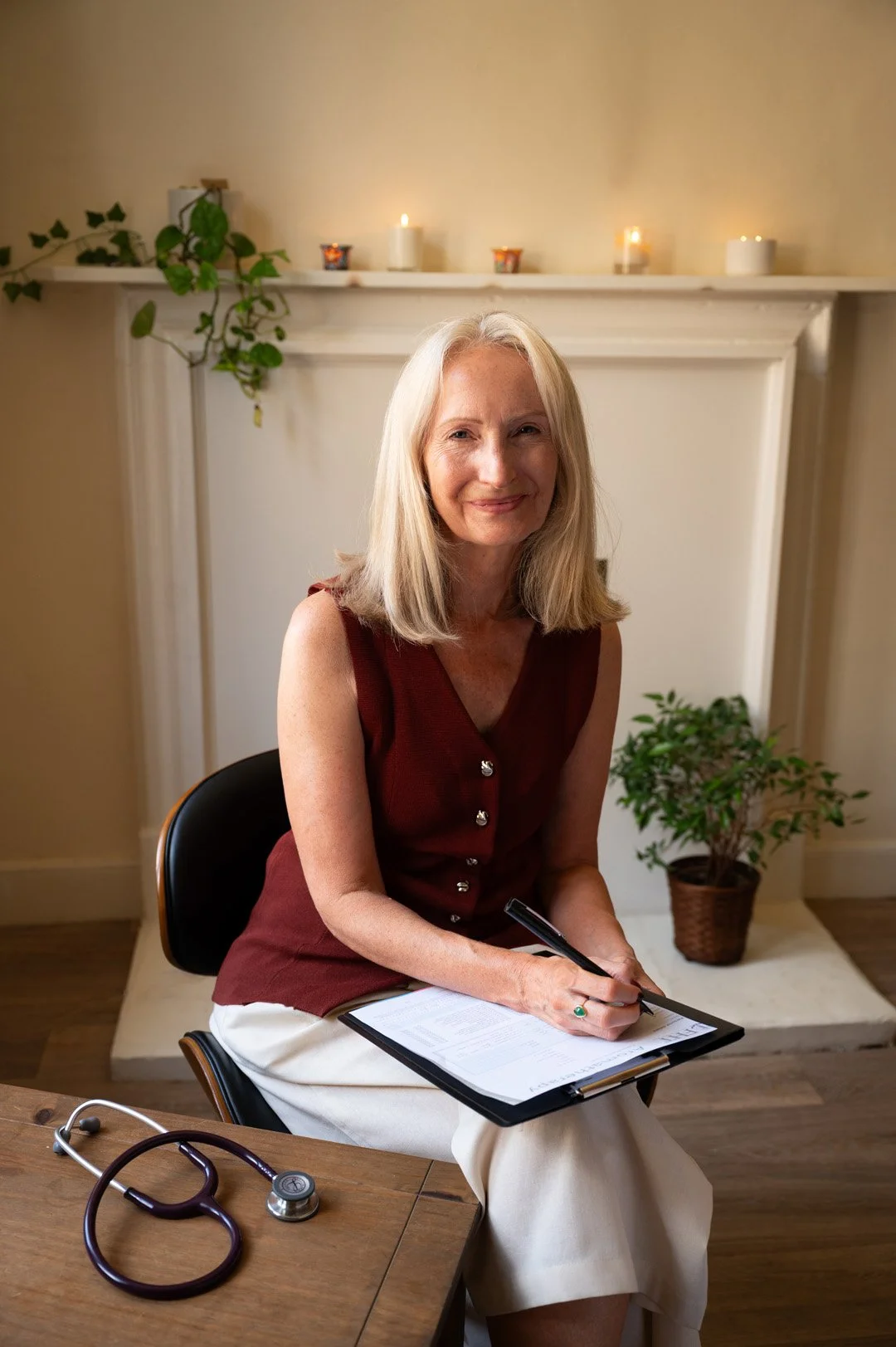 Julie smiling, sitting on a chair, holding a clipboard and pen, in a room with candles, plants, and a fireplace.
