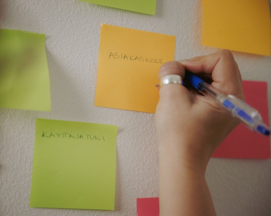 Käsi kirjoittamassa seinällä olevaan post -it -lappuun sanaa asiakaskokemus.