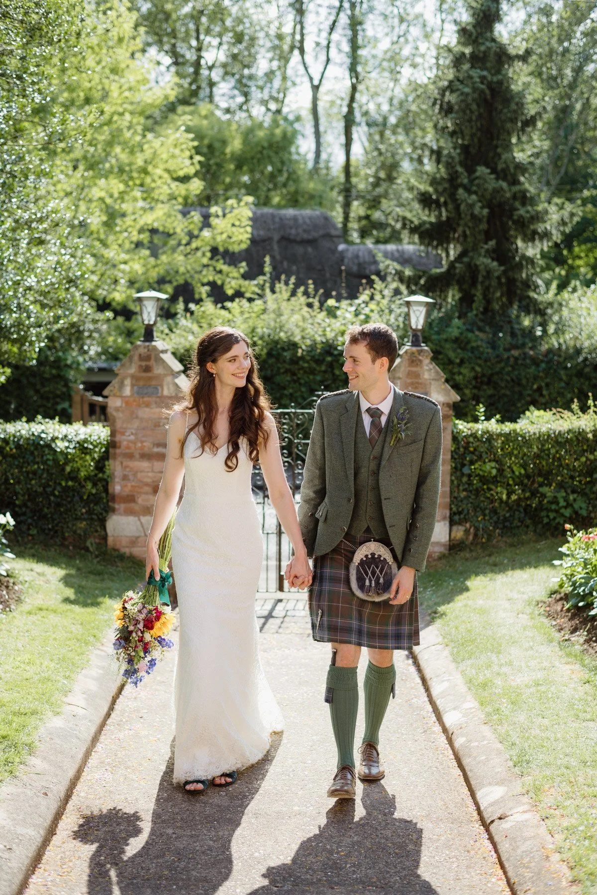 A bride in a white dress and a groom in traditional Scottish kilt walk hand-in-hand outdoors on a sunny day, smiling at each other, with a garden and stone gate in the background.