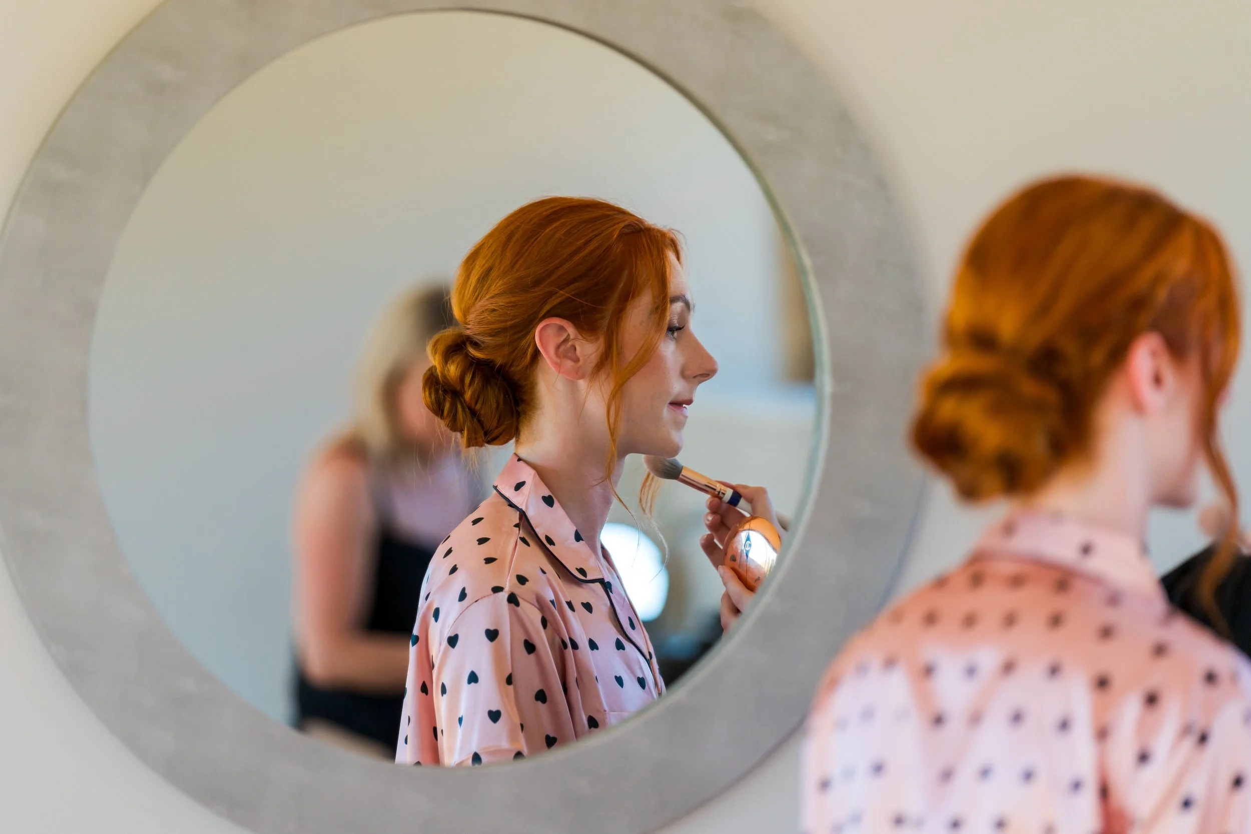A woman with red hair styled in a loose bun while getting her makeup done. She is wearing a pink pajama top with black hearts, and she is looking into a mirror as a makeup brush is applied to her face.