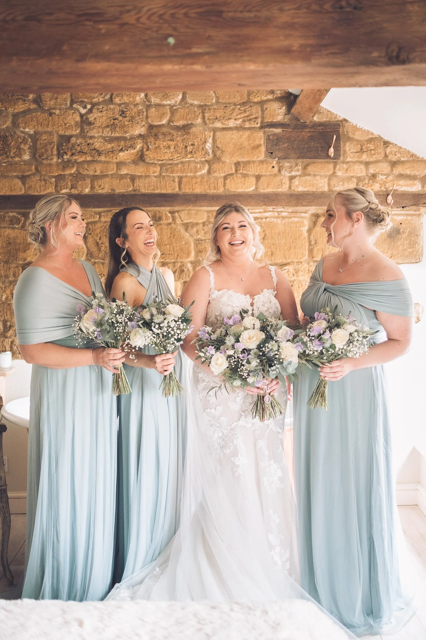A bride and three bridesmaids standing together, all holding bouquets of flowers, in a room with brick and wooden walls. The bride is in a white wedding dress, and the bridesmaids are in matching light blue gowns. They are smiling and appear joyful.