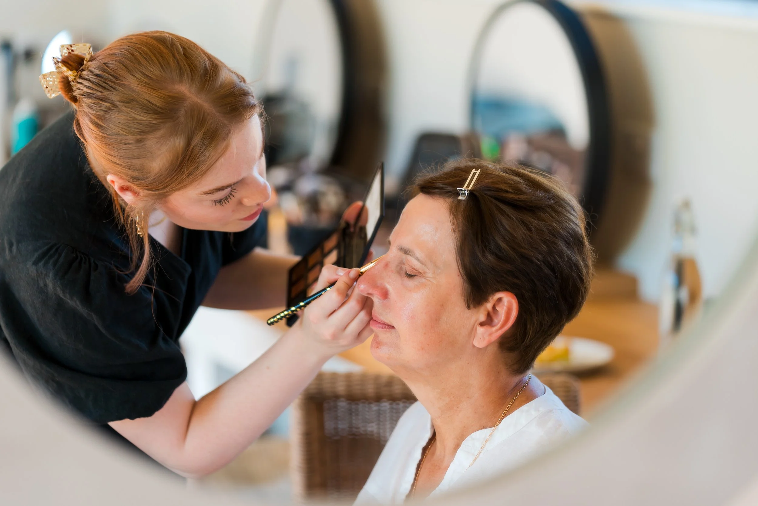 A woman applies makeup to a woman's face while looking into a mirror.