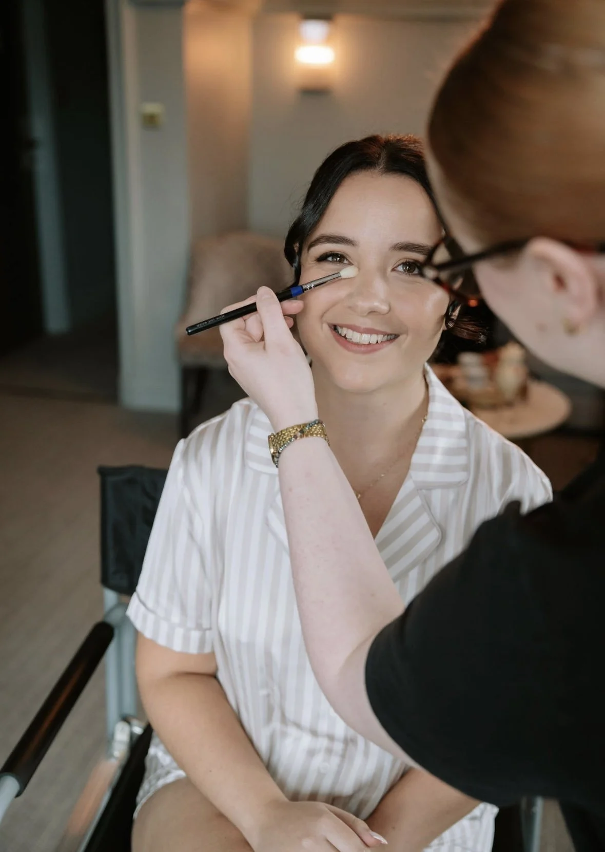 A woman sitting in a chair is having makeup applied by a makeup artist. The woman is smiling and wearing a striped shirt, with a gold bracelet on her wrist.