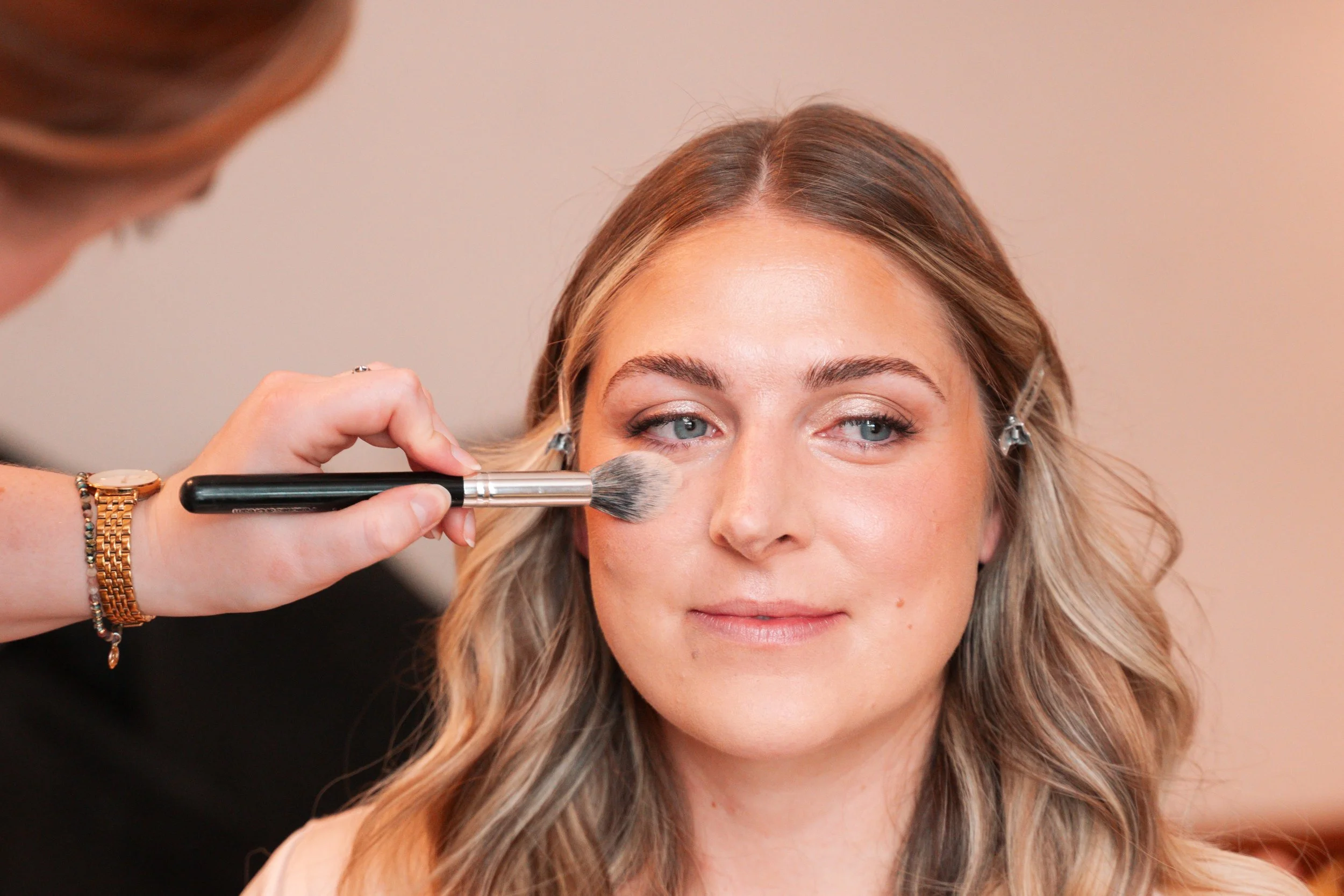 A woman with blonde hair sitting while a makeup artist applies blush to her cheek with a brush.