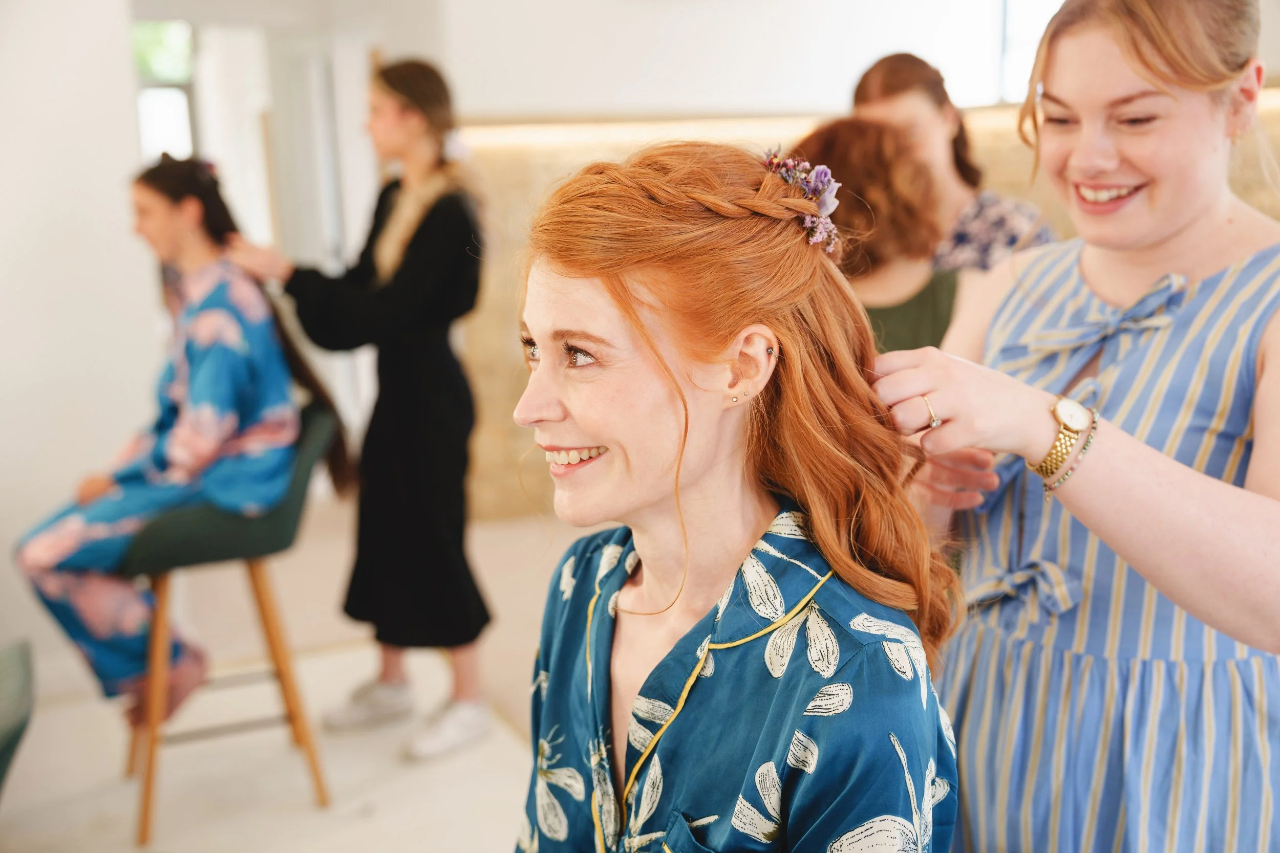 A woman with red hair is getting her hair styled by a stylist, who is smiling and dressed in a striped dress. Another woman is seated in the background, getting her hair done, while a stylist works on her hair. The setting appears to be a salon.