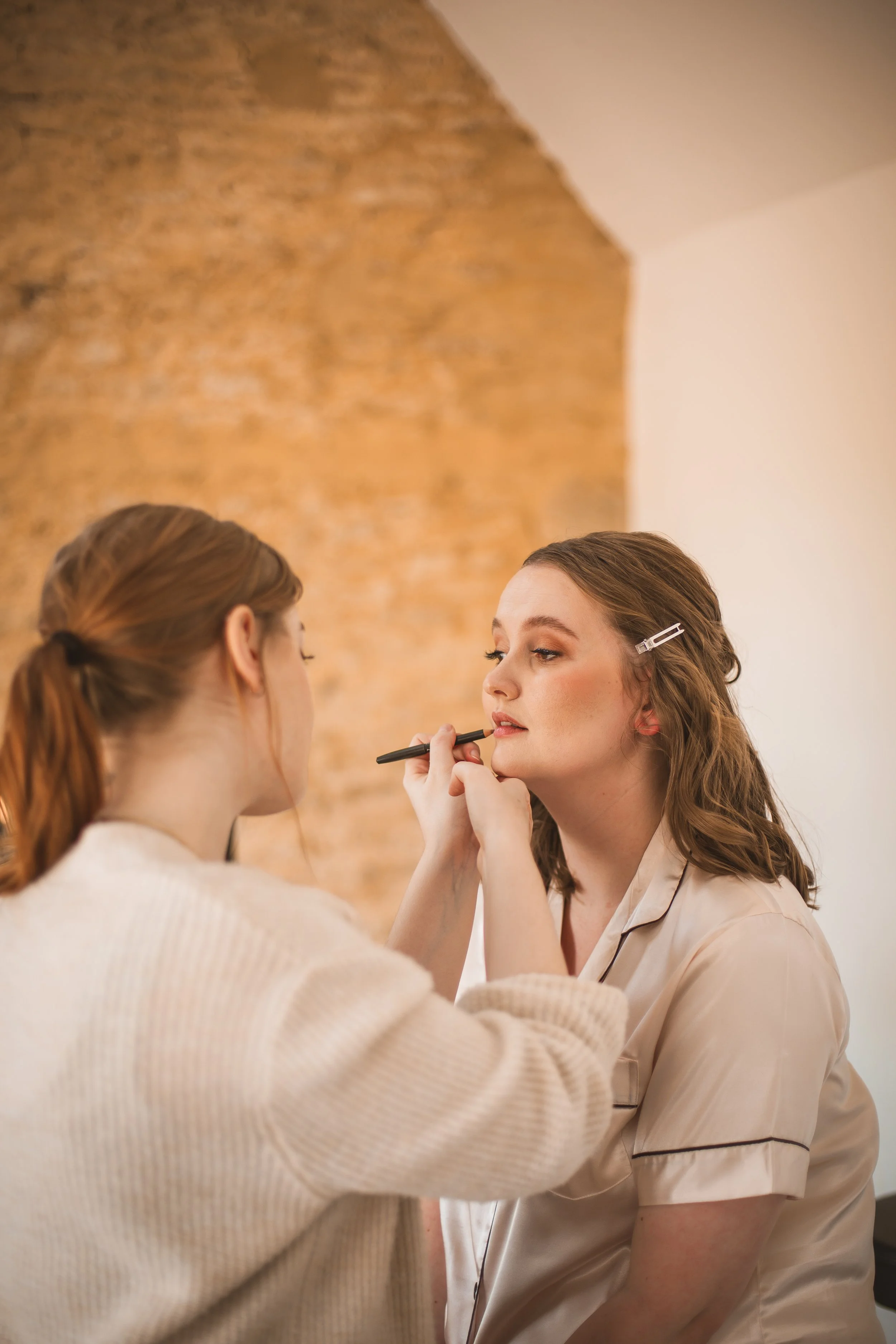 Makeup artist applying lipstick to a woman in a beige pajamas, with a brick wall background.