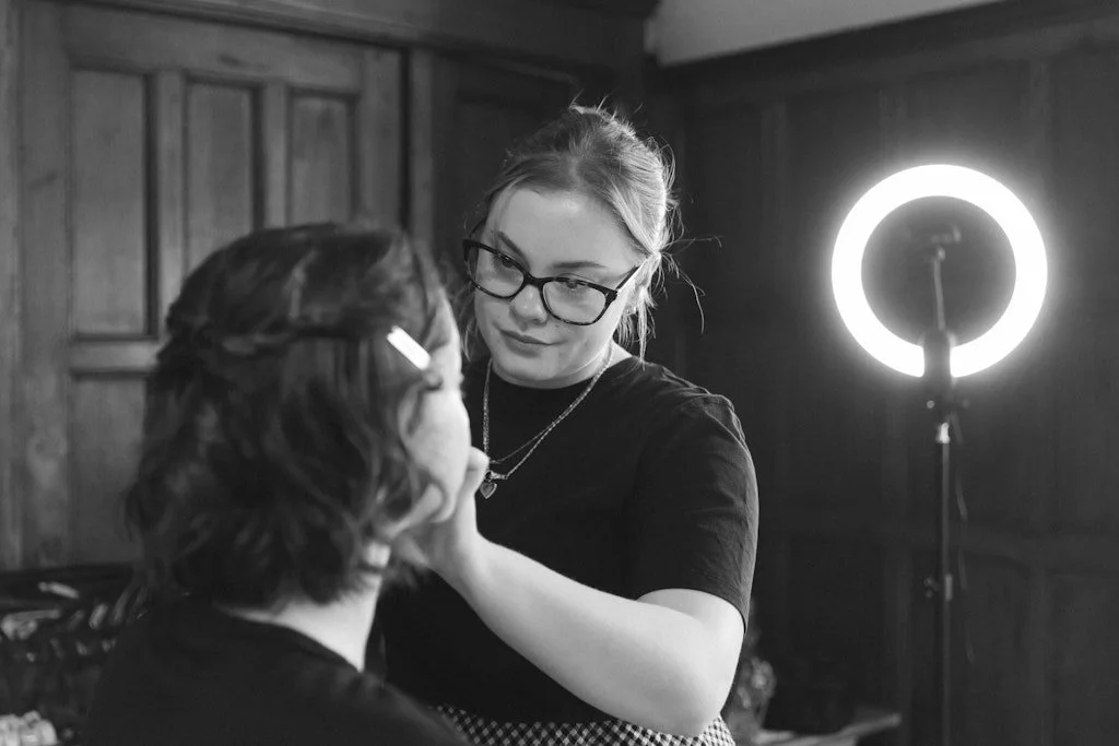 A makeup artist applying makeup to a woman with short curly hair, in a room with wooden walls and a ring light in the background.