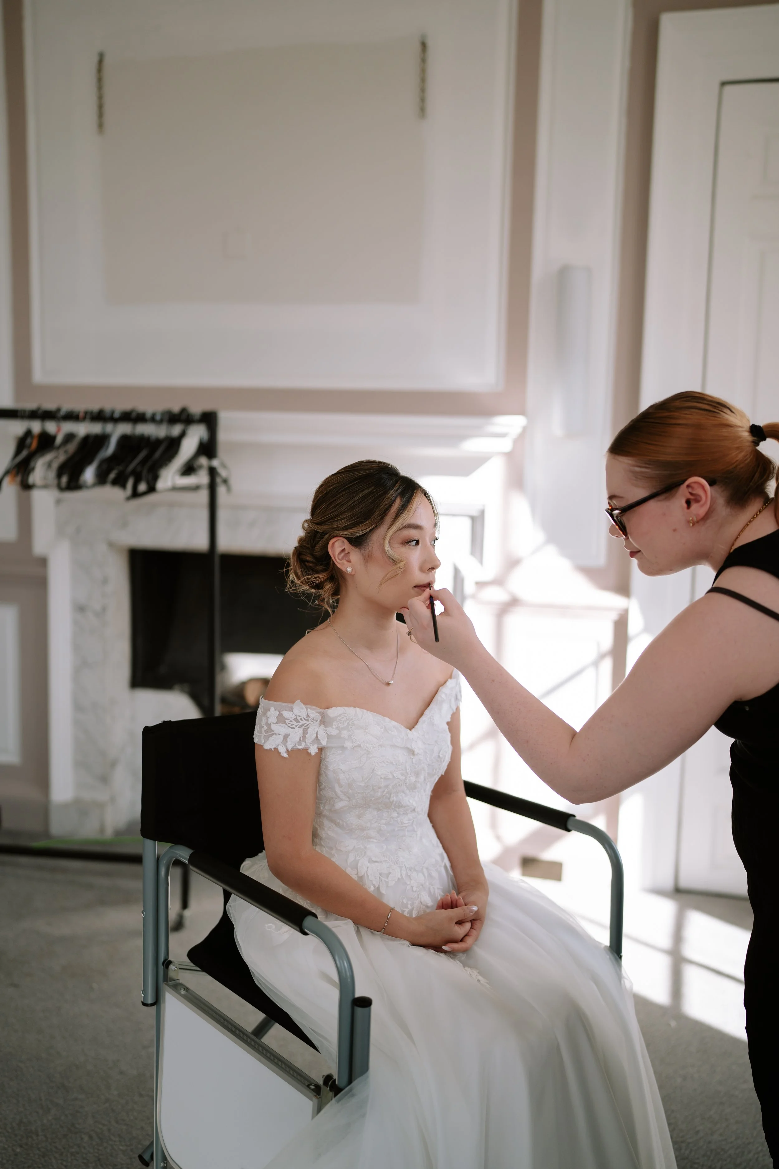 A bride with an off-shoulder wedding dress sitting in a chair while a makeup artist applies makeup to her face indoors.