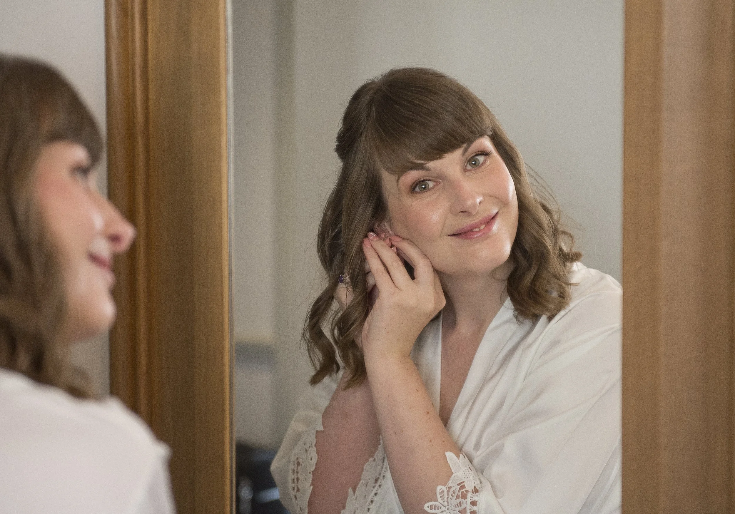 A woman with shoulder-length brown hair looking at herself in a mirror, smiling, while adjusting her earring.