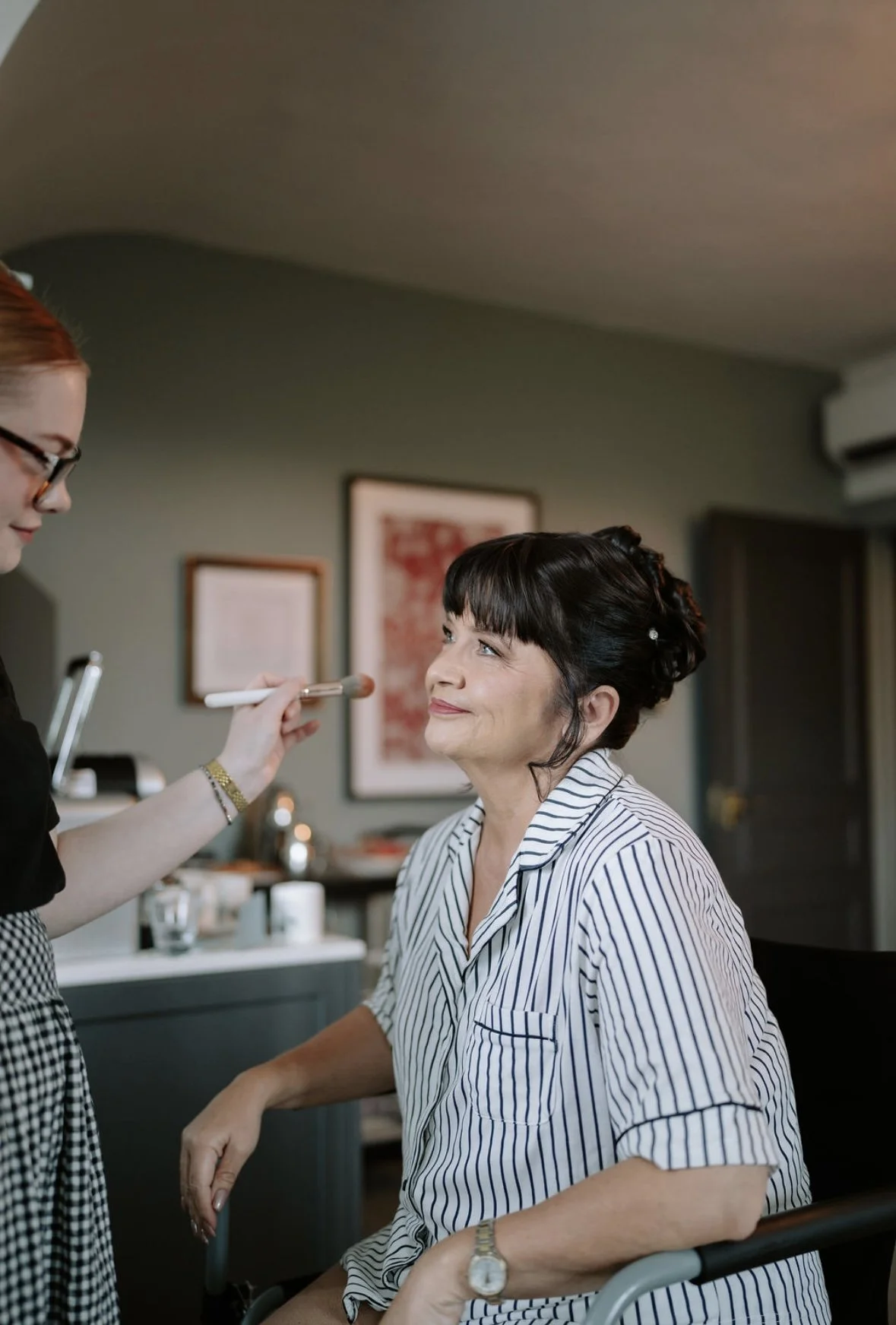 Woman in striped pajamas getting her makeup done by a makeup artist in a cozy room.