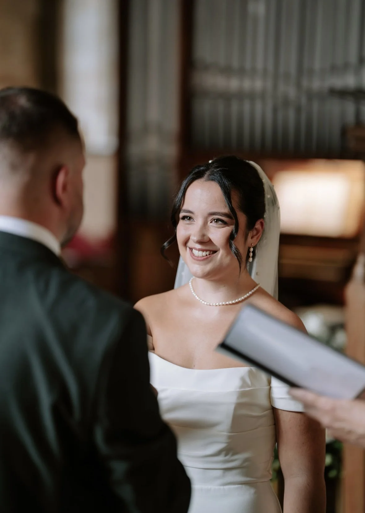 A bride with black hair, wearing a white dress, pearl necklace, and earrings, smiling during her wedding ceremony.