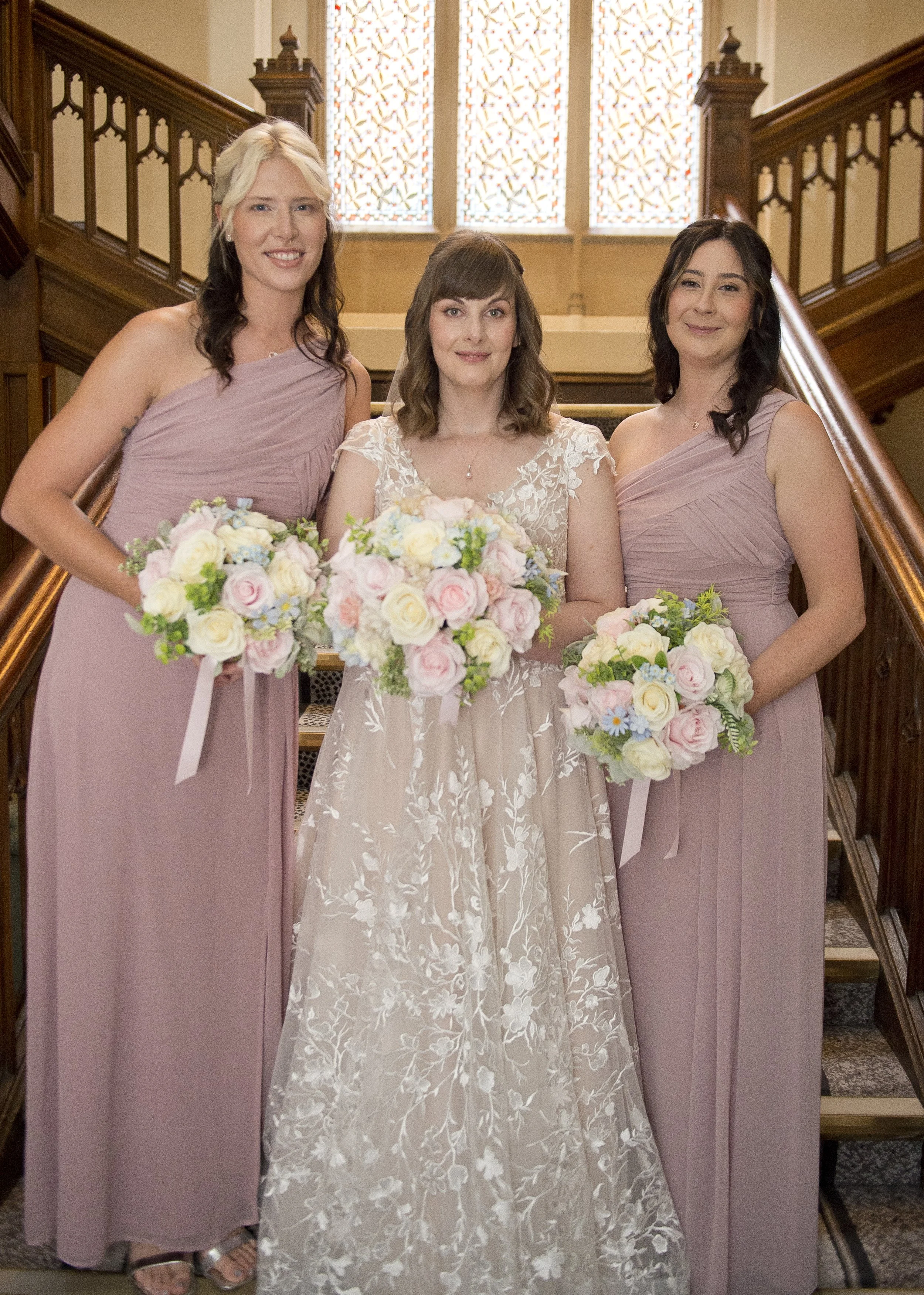 A bride in a lace wedding gown standing between two bridesmaids in mauve dresses, all holding bouquets of flowers, inside a historic building with wooden stairs and stained glass windows.